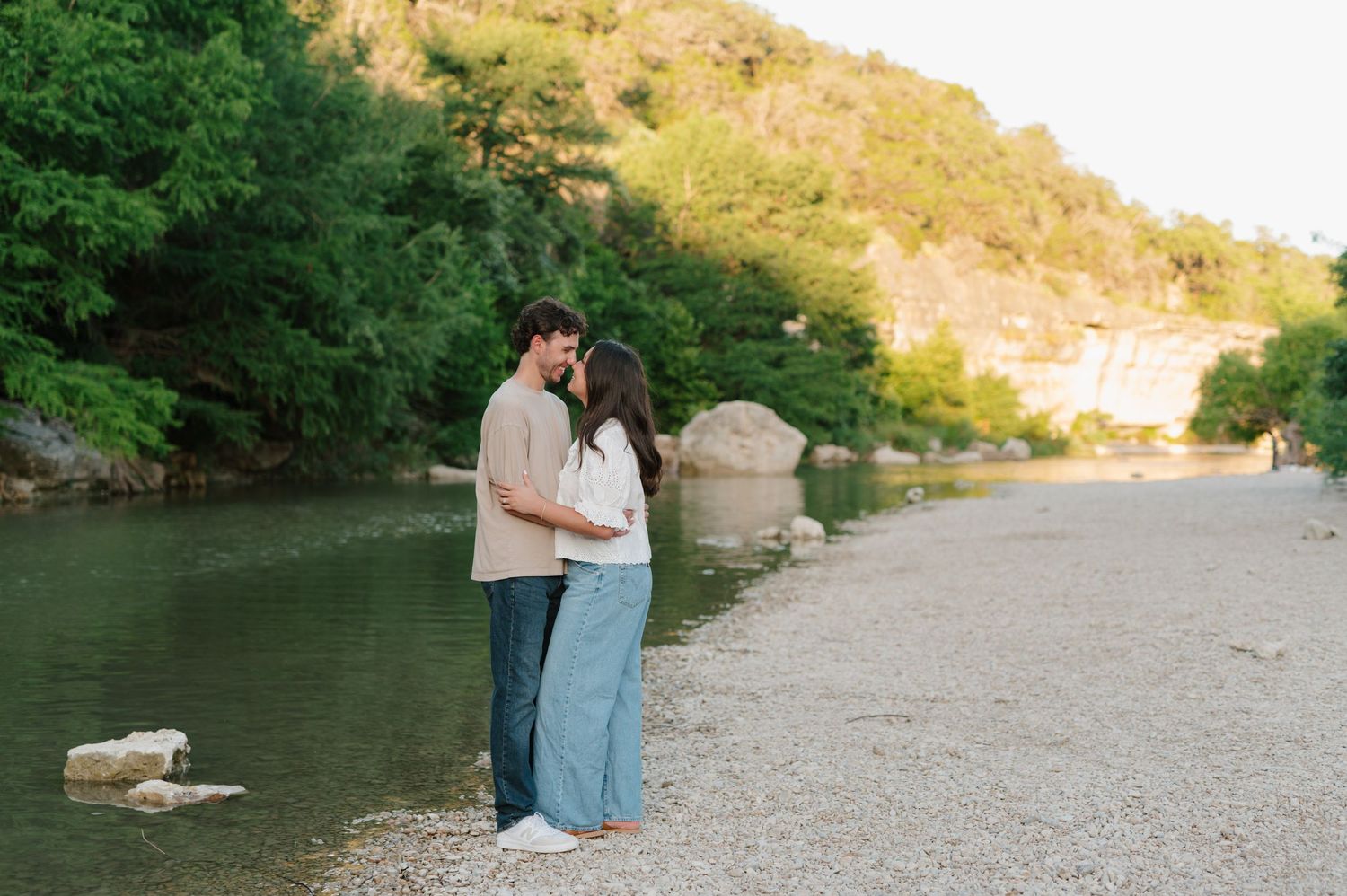Couple shares intimate moment along riverbank with hills in background.