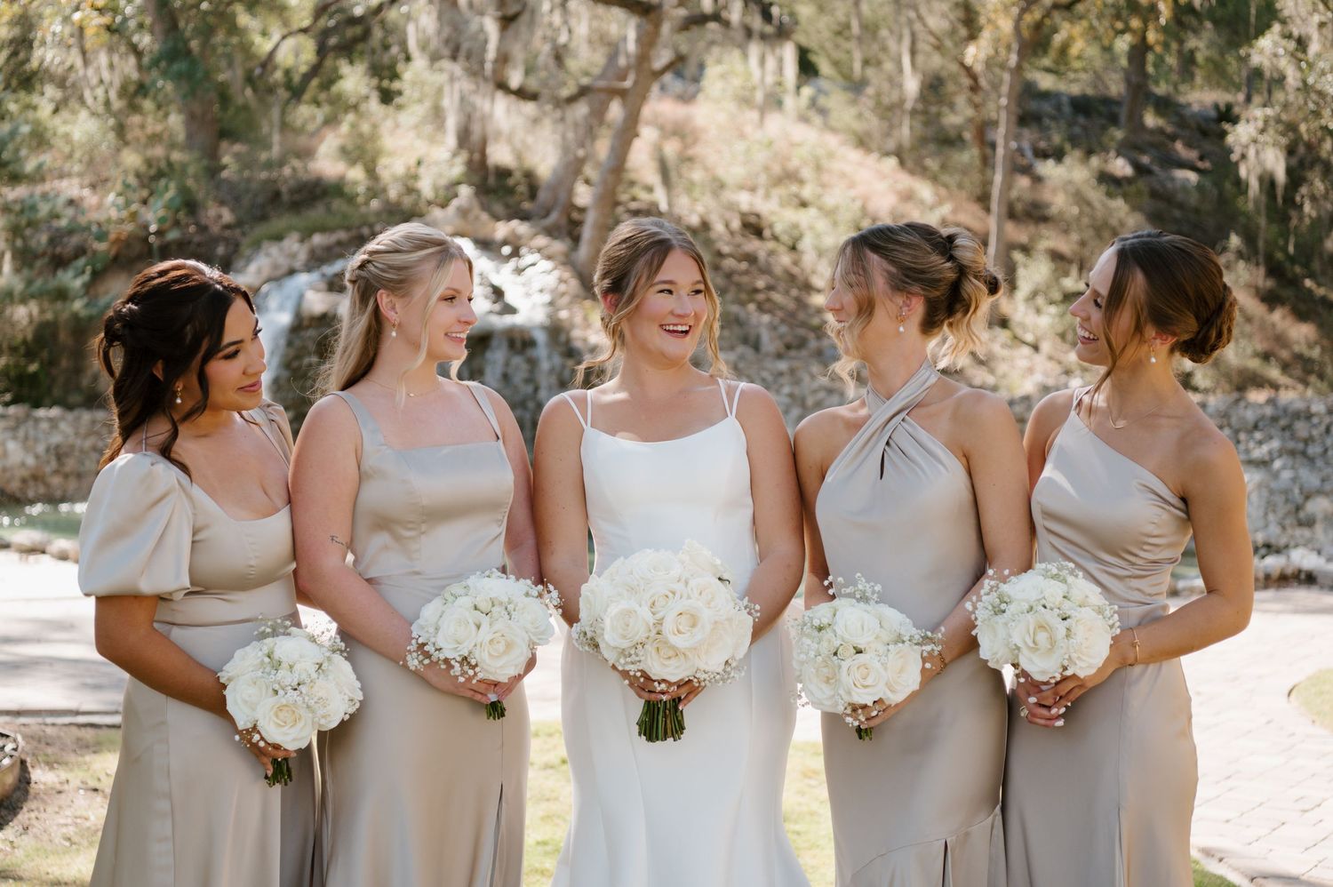 Wedding party wearing light gray bridesmaid dresses holding white bouquets pose together outdoors.
