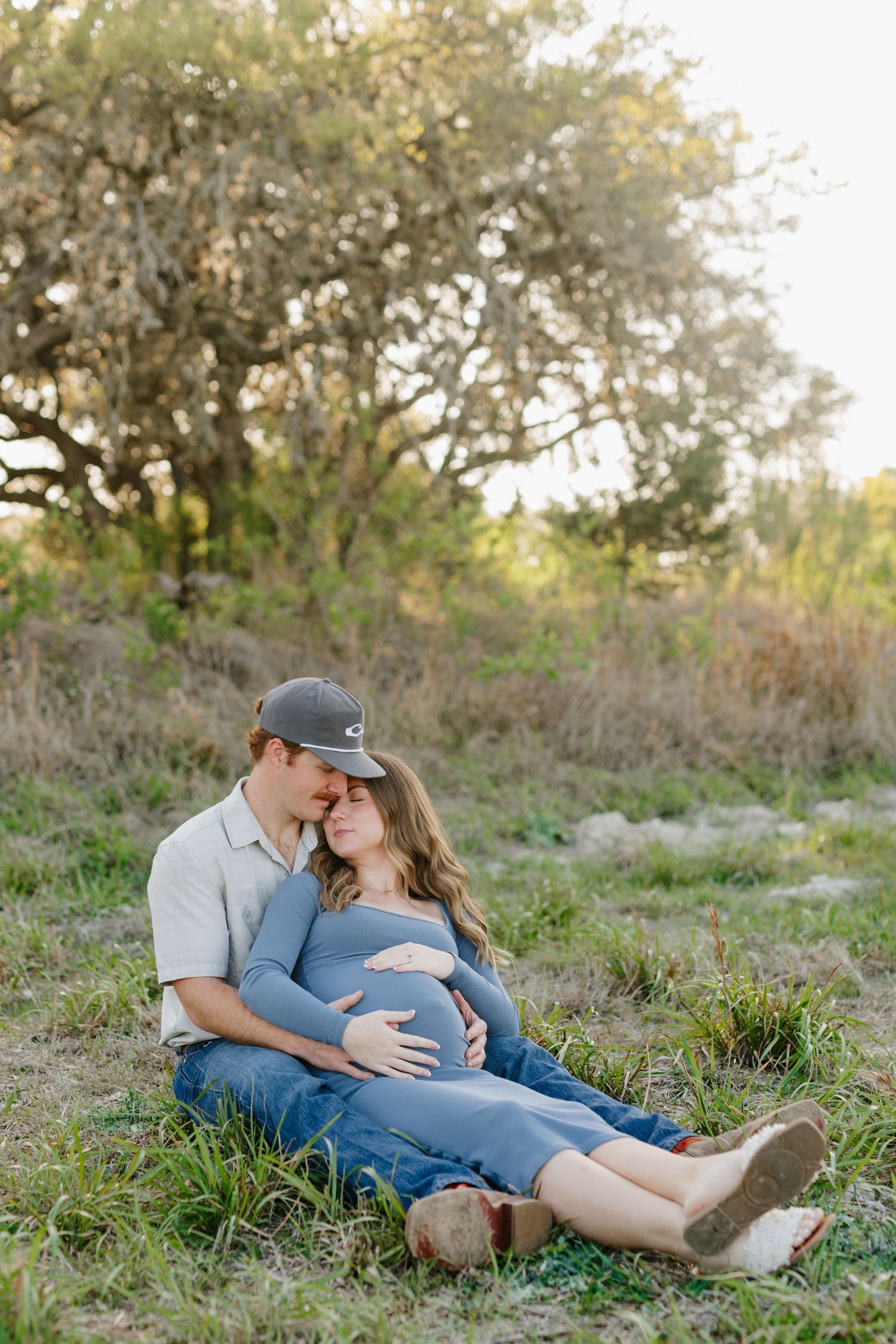 A couple relaxes on grass beneath a tree during a maternity photo session in golden sunlight.