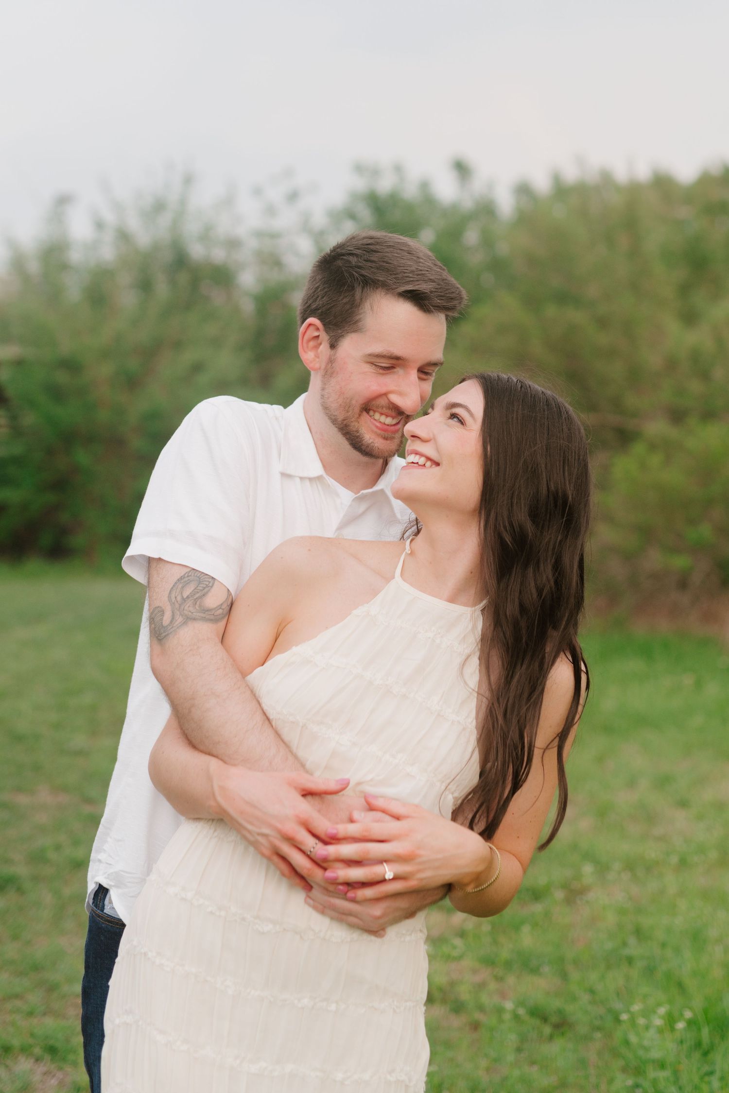 A couple shares a romantic moment outdoors in a field wearing light colored outfits during an engagement photo session.
