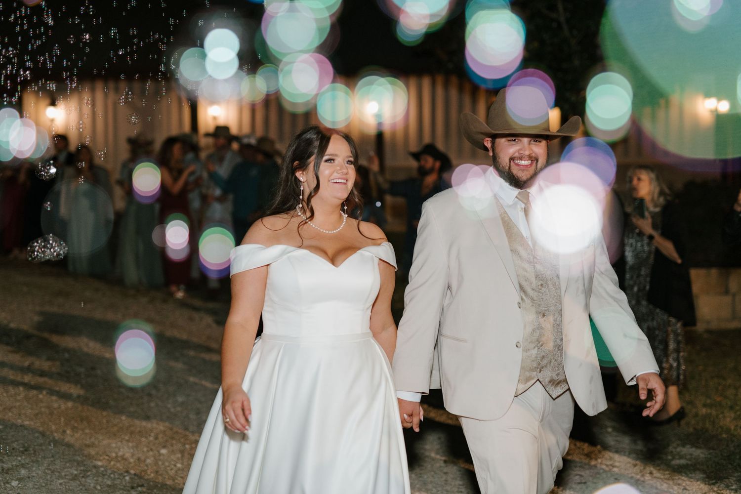 A couple in wedding attire walks together at night under glowing lights creating a dreamy bokeh effect.