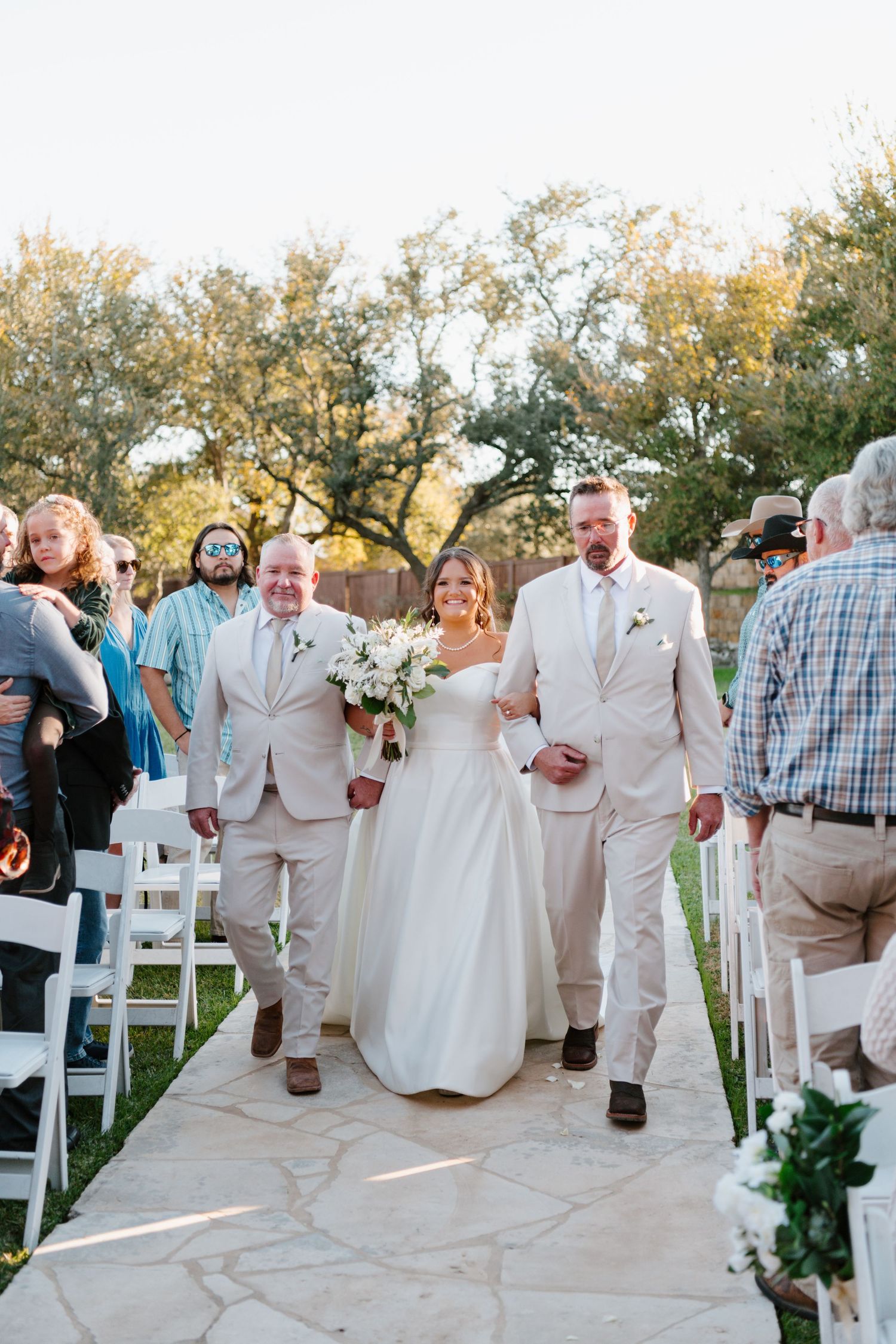 Happy couple walking down the stone aisle after outdoor wedding ceremony with guests looking on among trees.