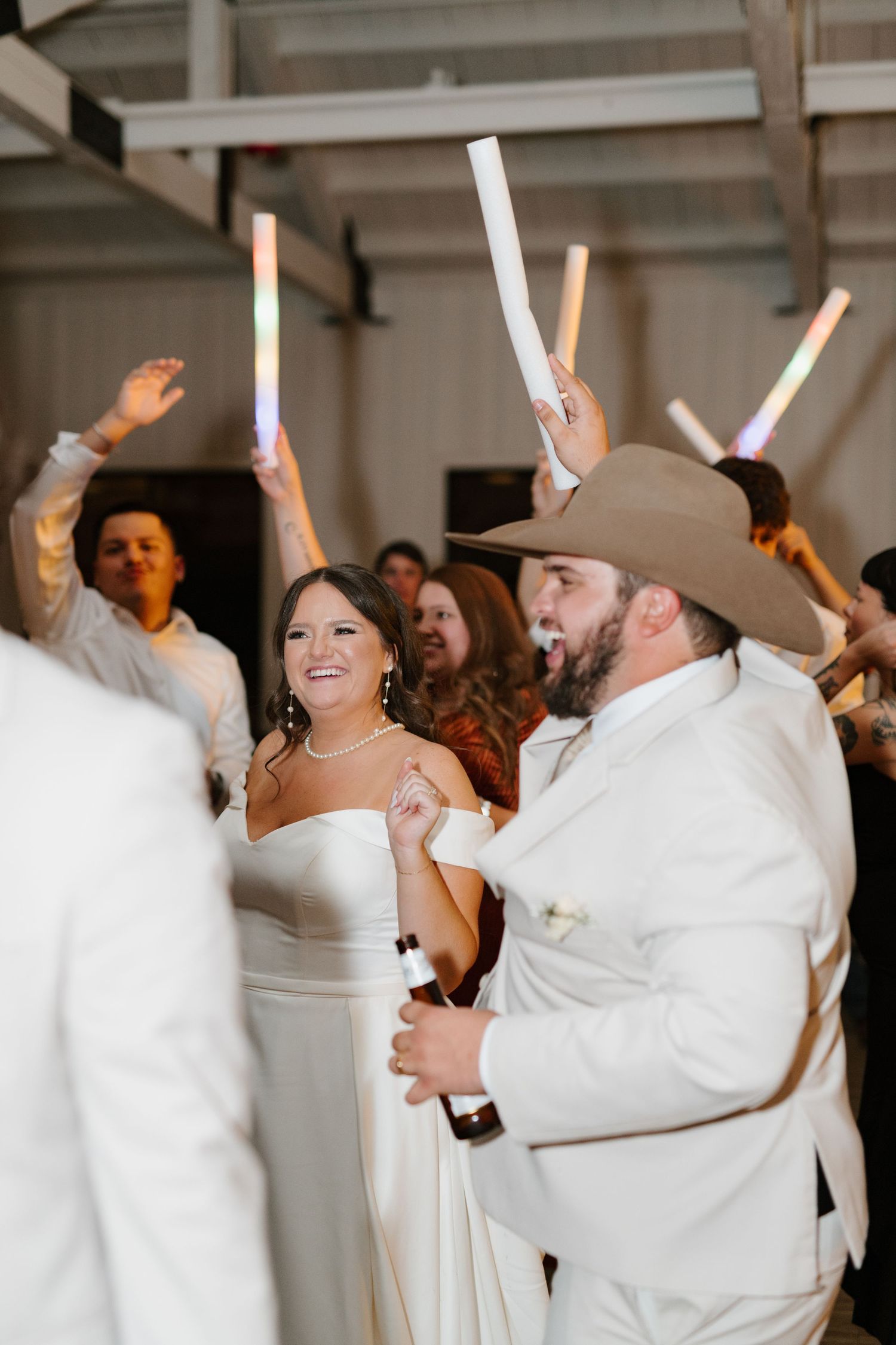 Wedding celebration with people dancing and holding glowing foam sticks while dressed in white attire.