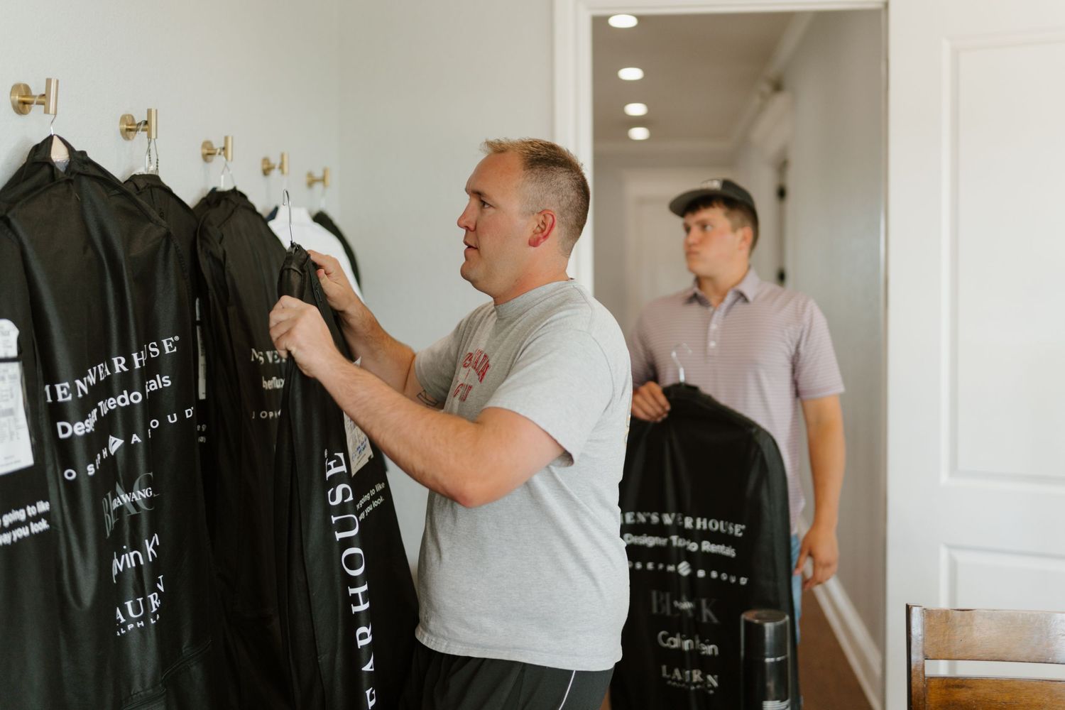 Someone inspects black jackets hanging on a clothing rack while another person stands in the background of a bright hallway.