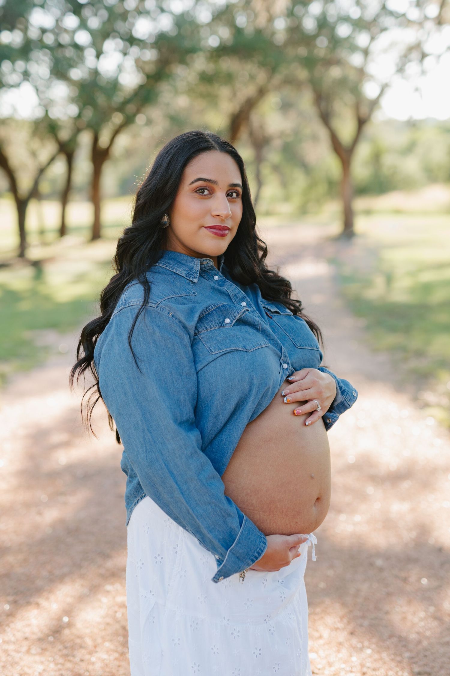 Expecting mother in denim jacket cradles her baby bump while standing in a sunlit outdoor setting.