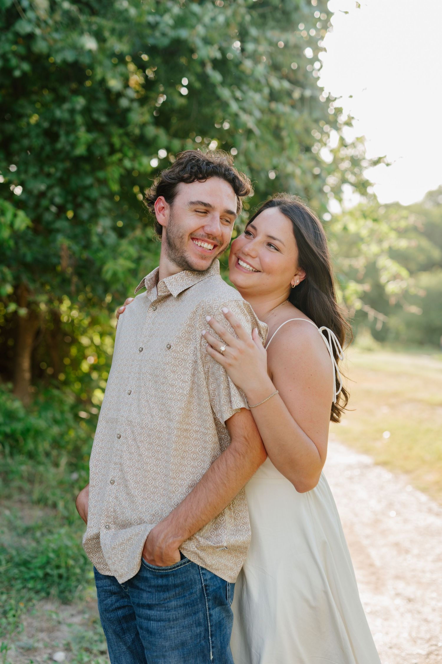 A couple in casual attire embraces while laughing together in a sunlit outdoor setting with green foliage.