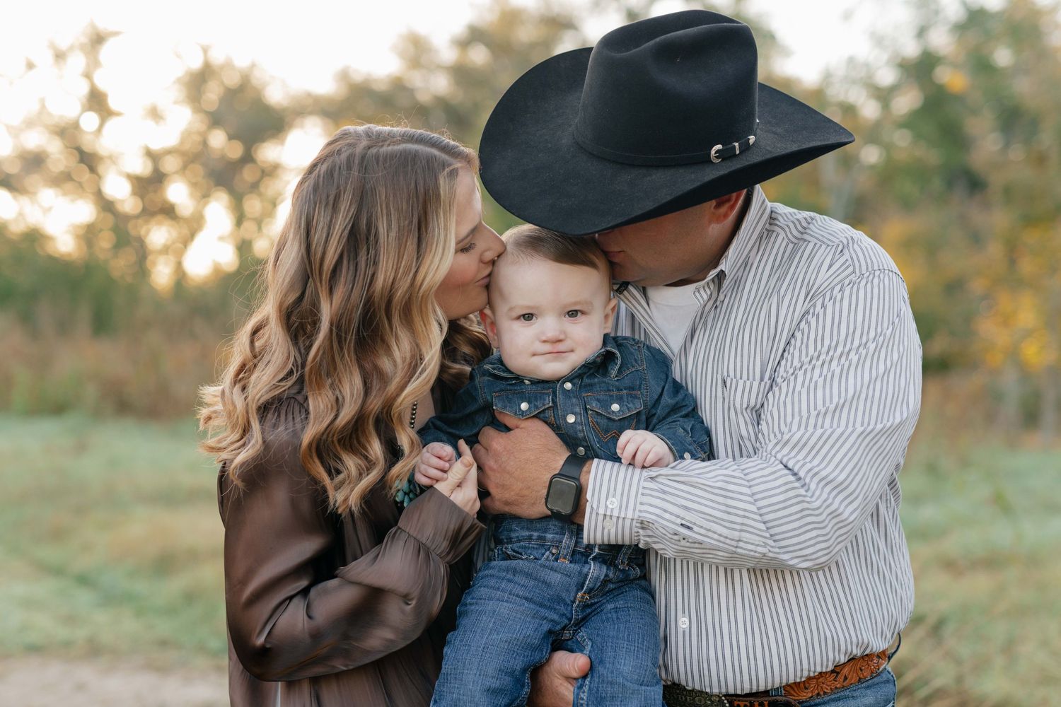 A family shares a tender moment outdoors at sunset in western-style clothing with a black cowboy hat.