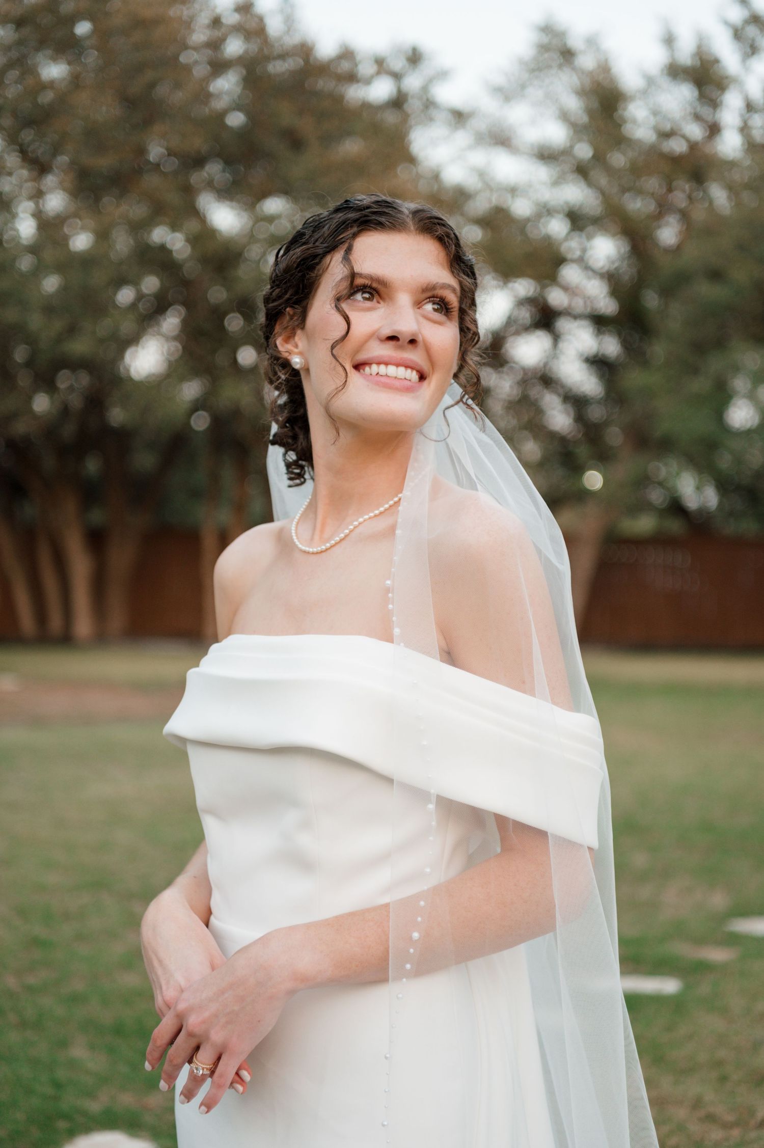 A joyful bride in an elegant off-shoulder wedding dress and veil poses outdoors during a photo session.