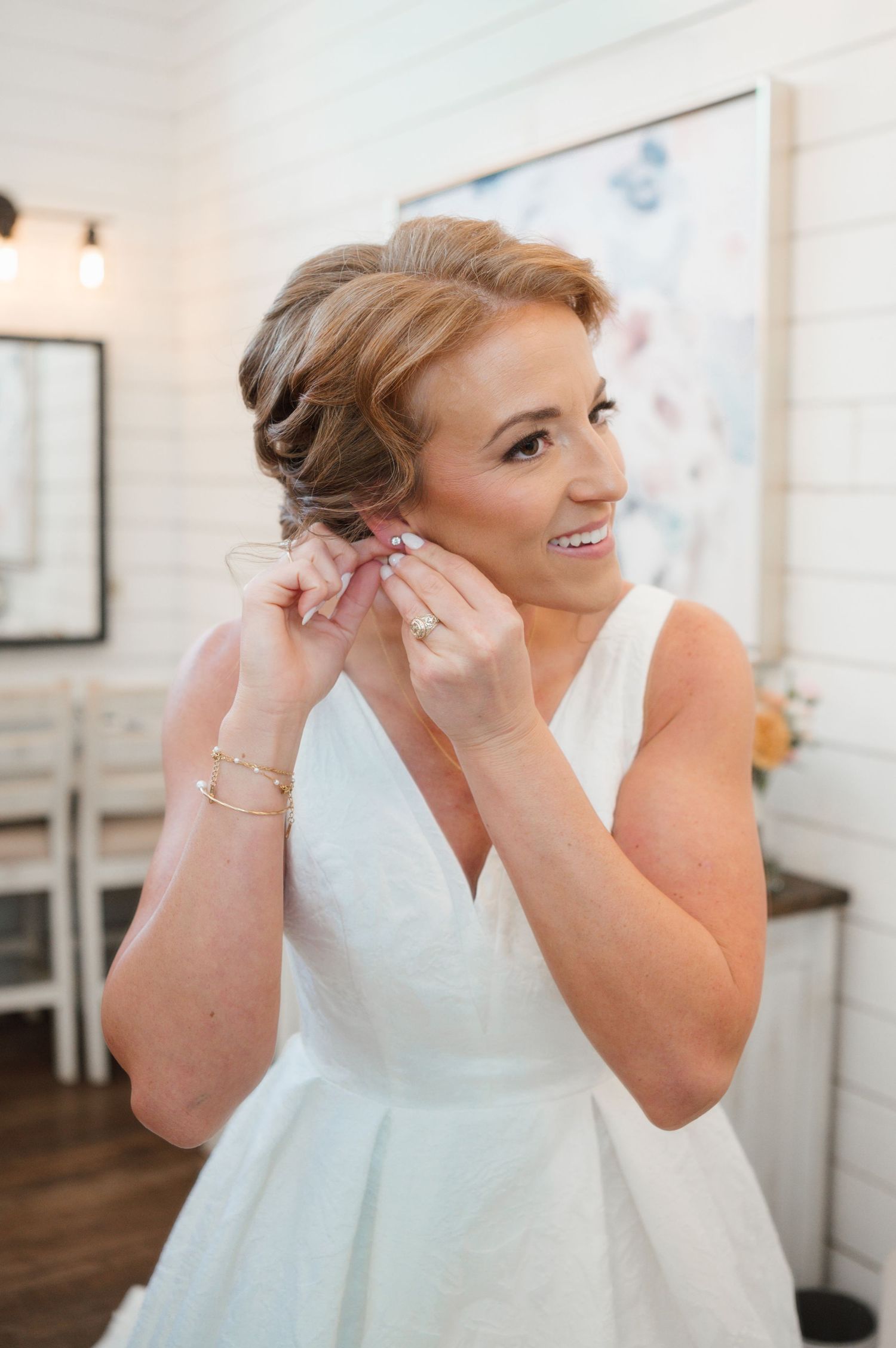 A bride in a classic white sleeveless wedding dress puts on earrings while smiling in a brightly lit room.