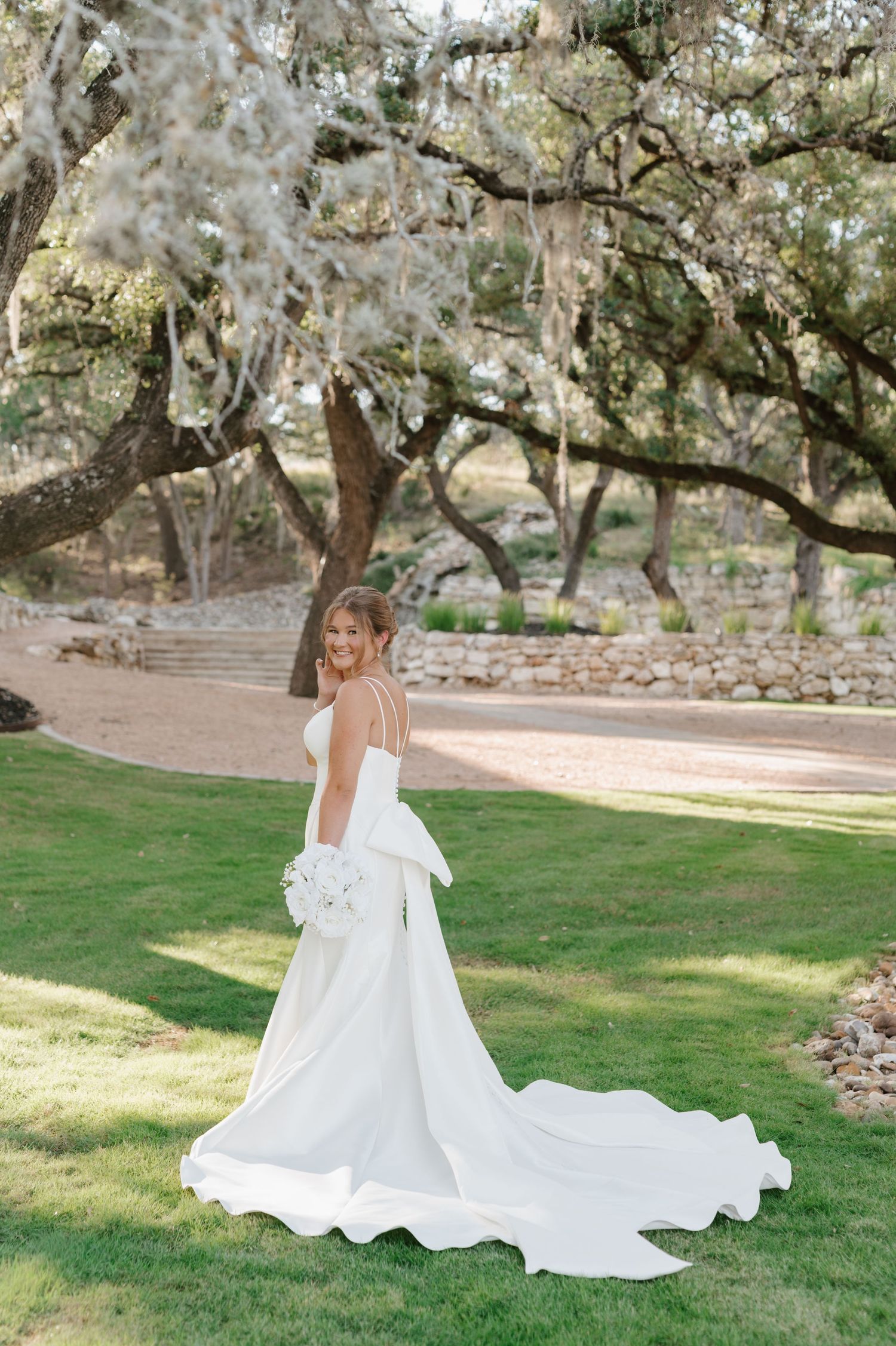 Bride in flowing white gown poses under blooming trees in garden setting.