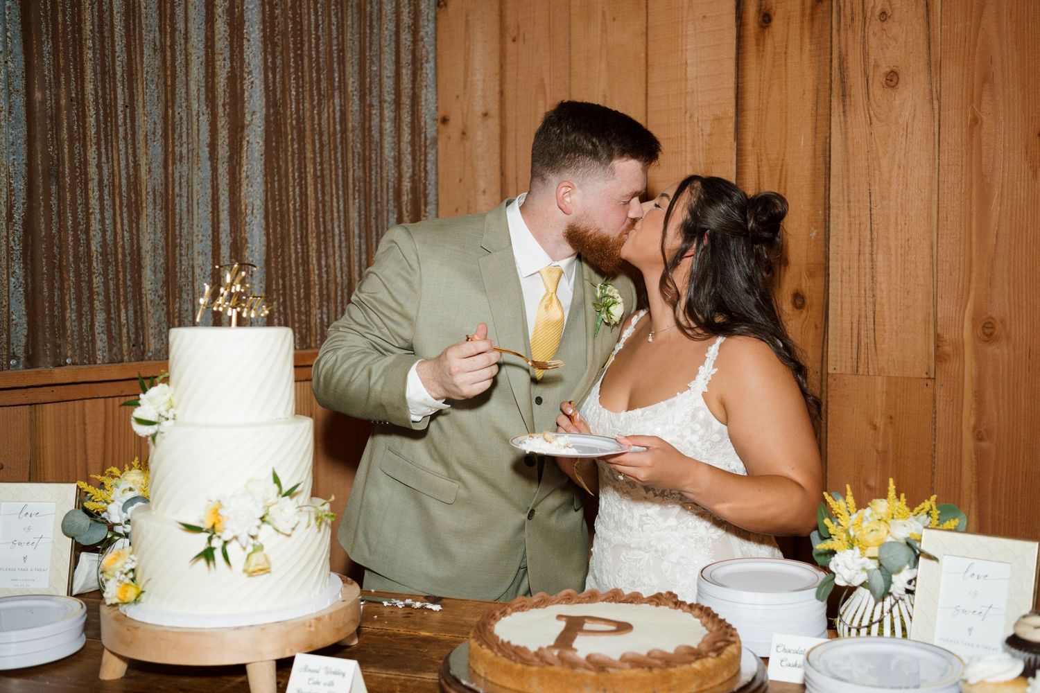 Bride and groom share a kiss while cutting their white floral wedding cake at their rustic reception.