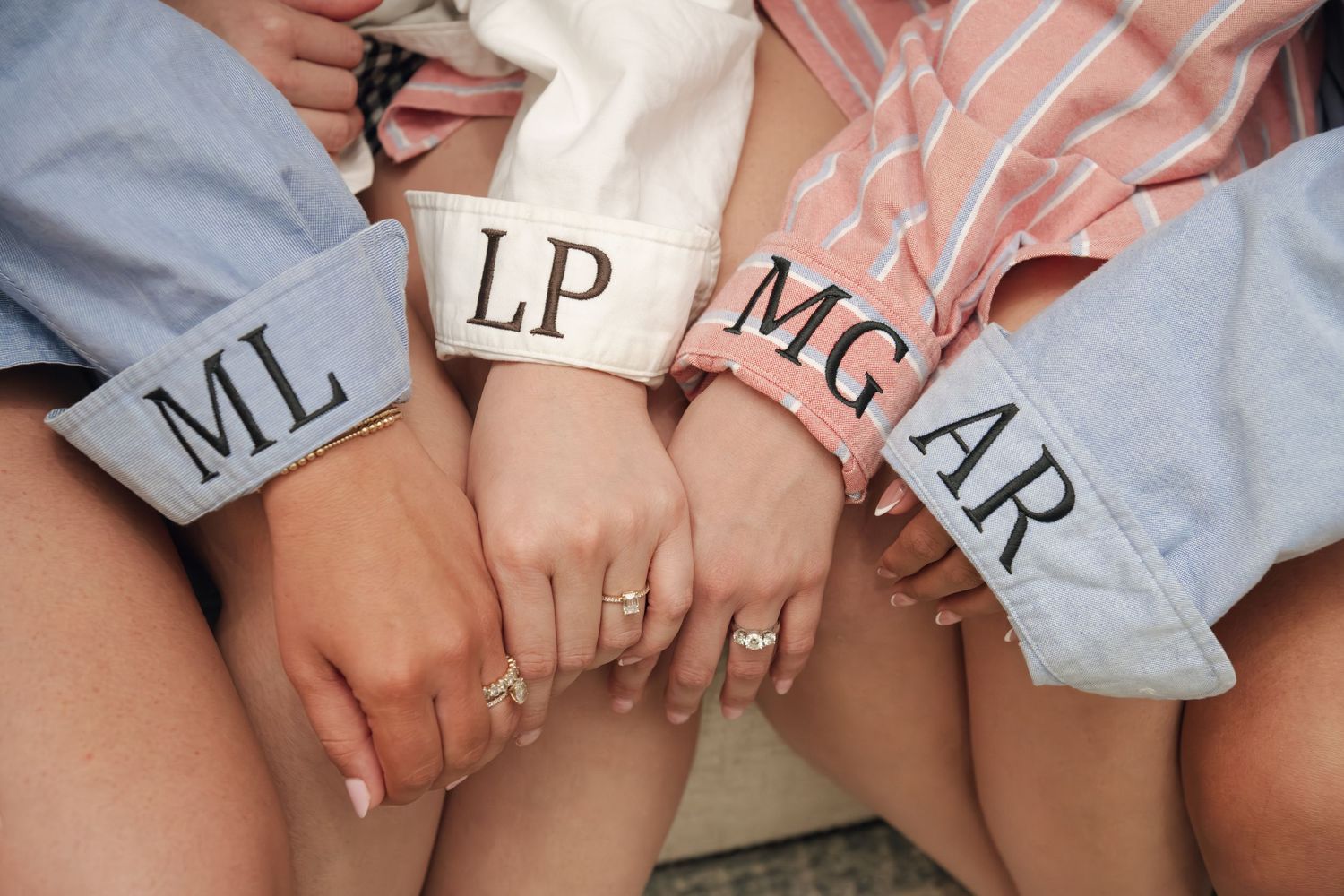 Four bridesmaids display monogrammed shirt cuffs with initials ML, LP, MG, and AR while wearing delicate gold rings.
