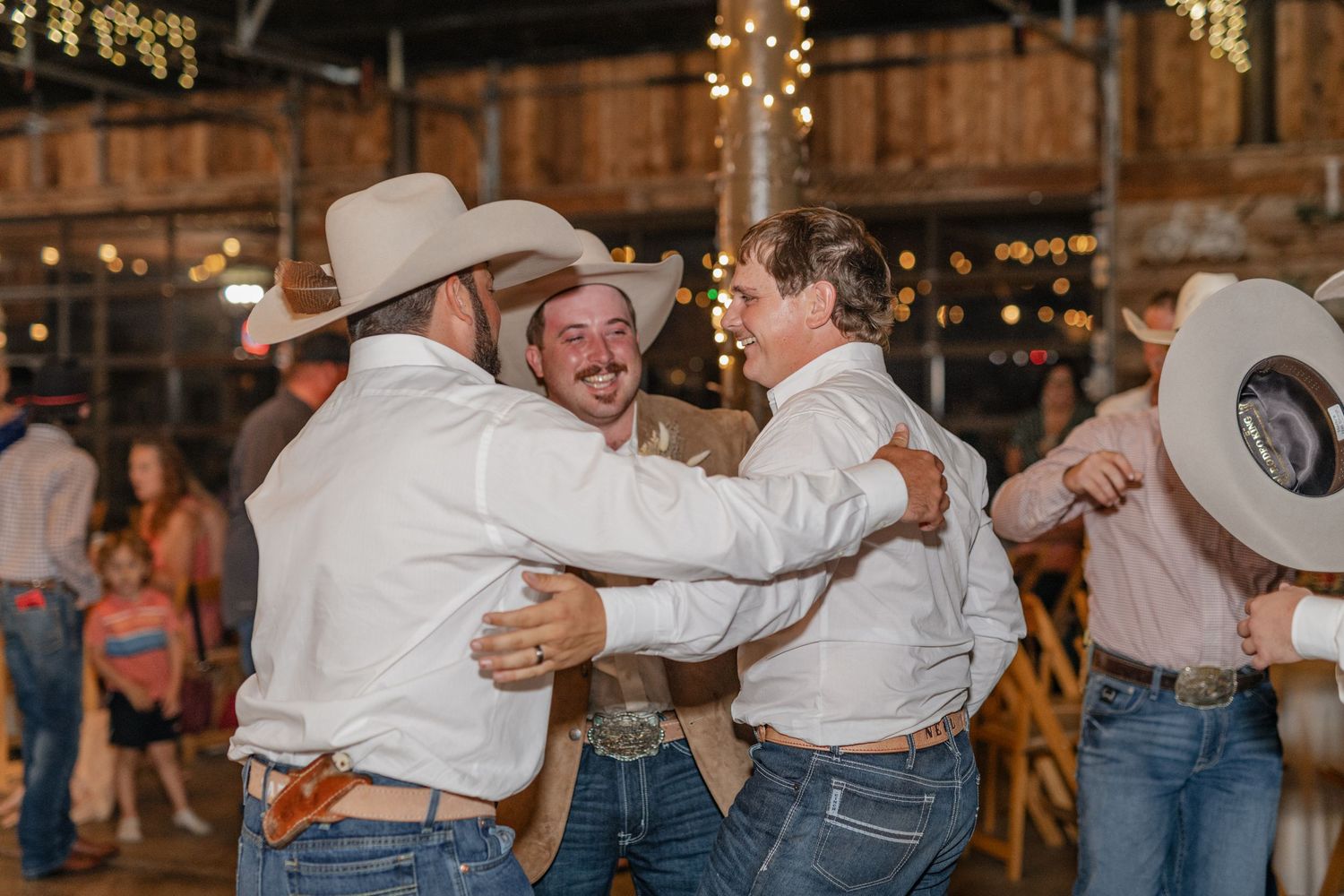 Men in cowboy hats share laughs and hugs during wedding reception in rustic barn venue.