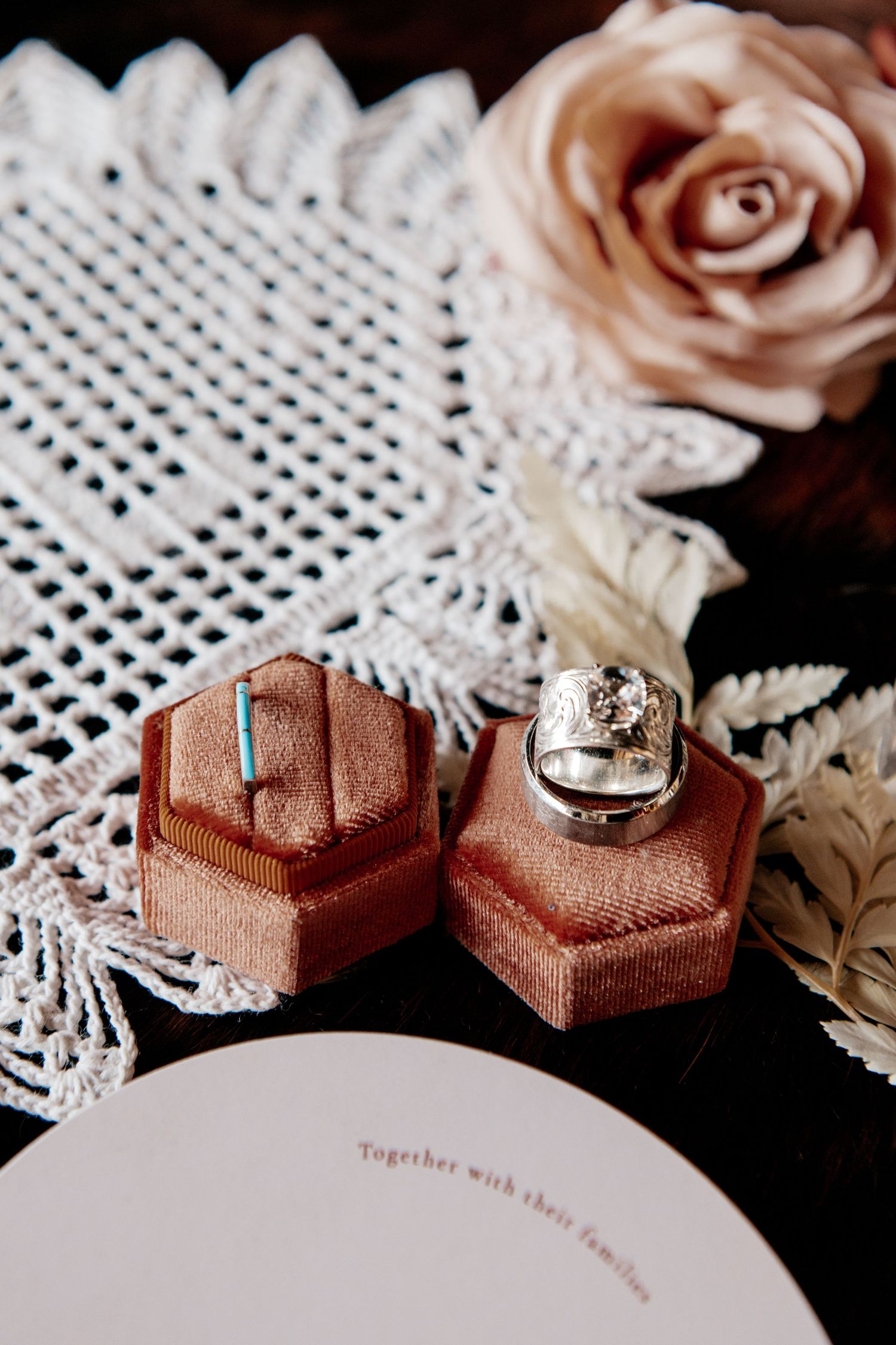 Wedding rings displayed in hexagonal velvet ring boxes on vintage lace with a blush rose accent.