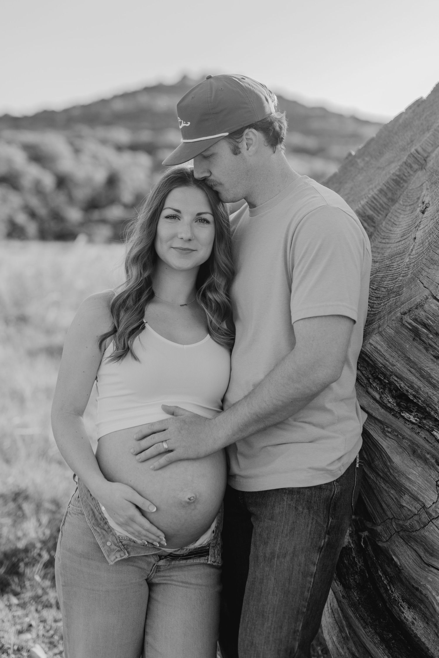 Black and white maternity portrait of couple in casual attire posed against wooden backdrop outdoors.