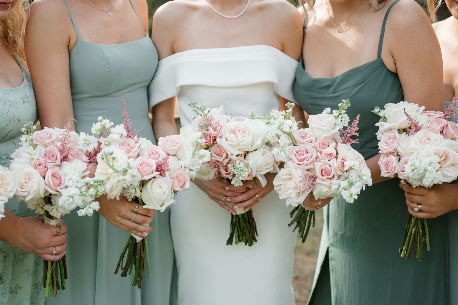 Bridesmaids in sage green dresses hold matching pink and white floral bouquets next to bride in white gown.