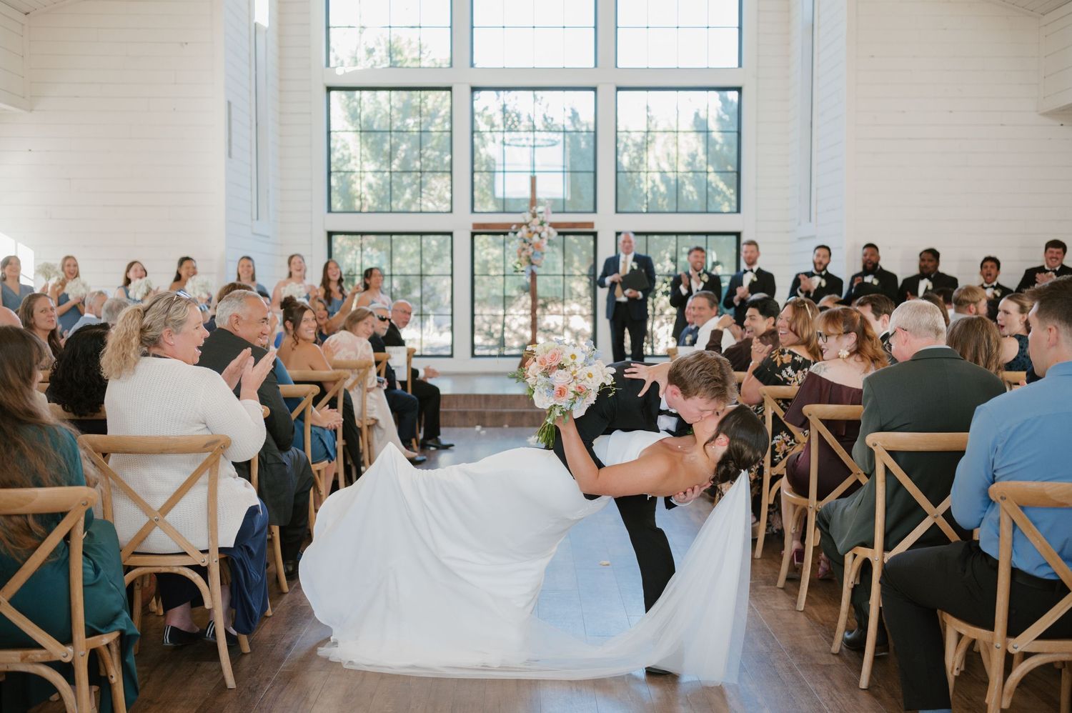 Wedding ceremony guests seated in rows of wooden chairs inside a bright sunlit venue with large windows.