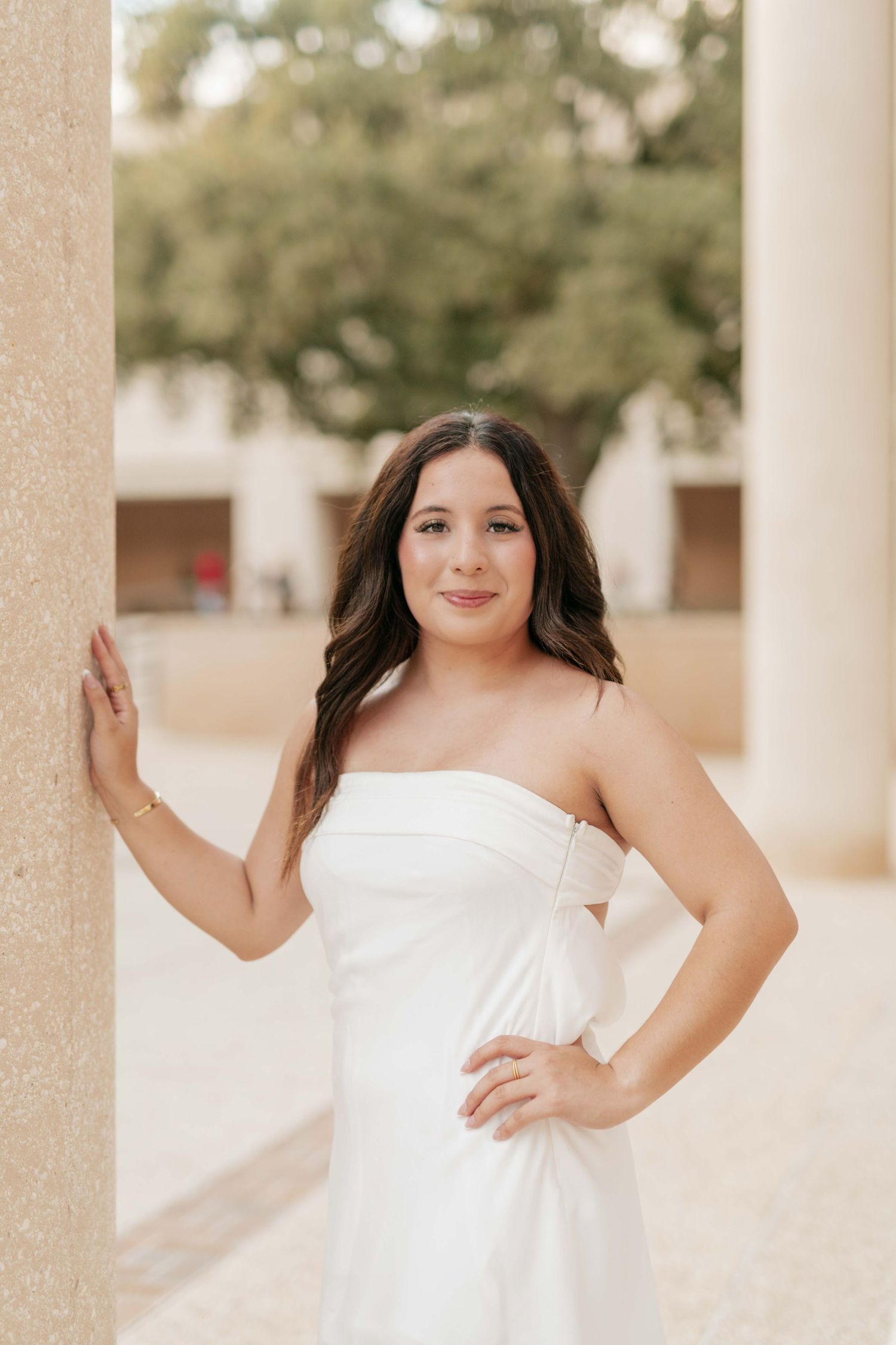 Graduate in white dress poses confidently against stone pillar.