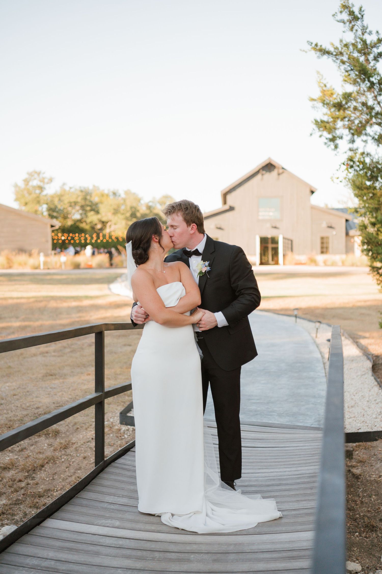 A couple in formal wedding attire shares romantic kisses on a wooden deck at sunset.