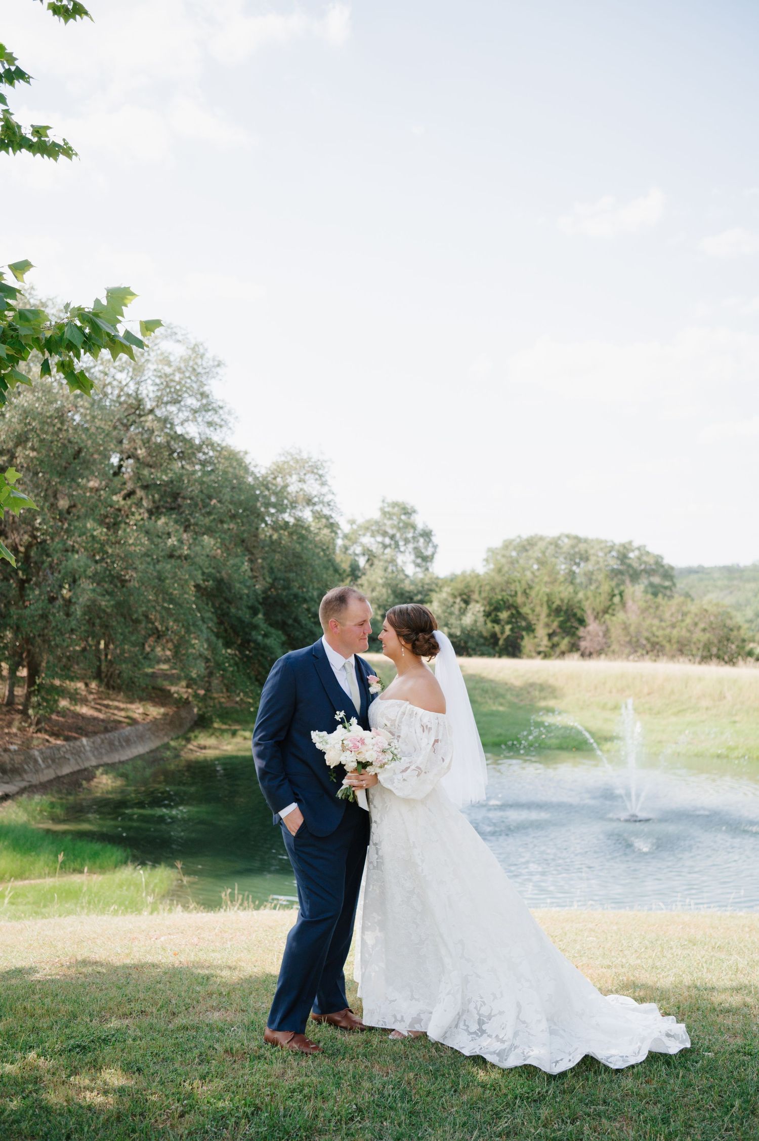 Elegant wedding couple photographed by a lake with fountain in the background.