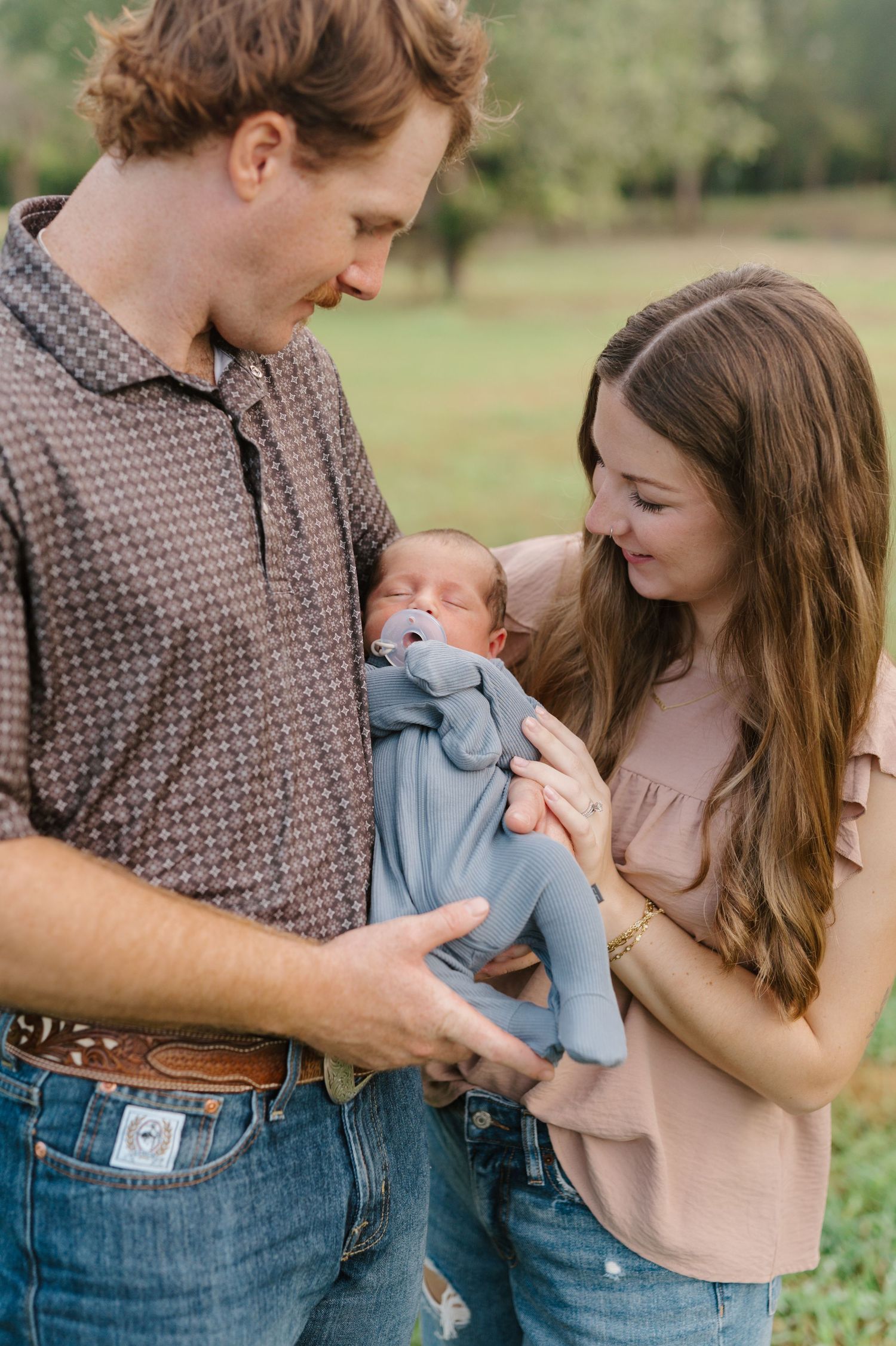 A young couple embraces tenderly while holding a small baby wrapped in blue in an outdoor setting.