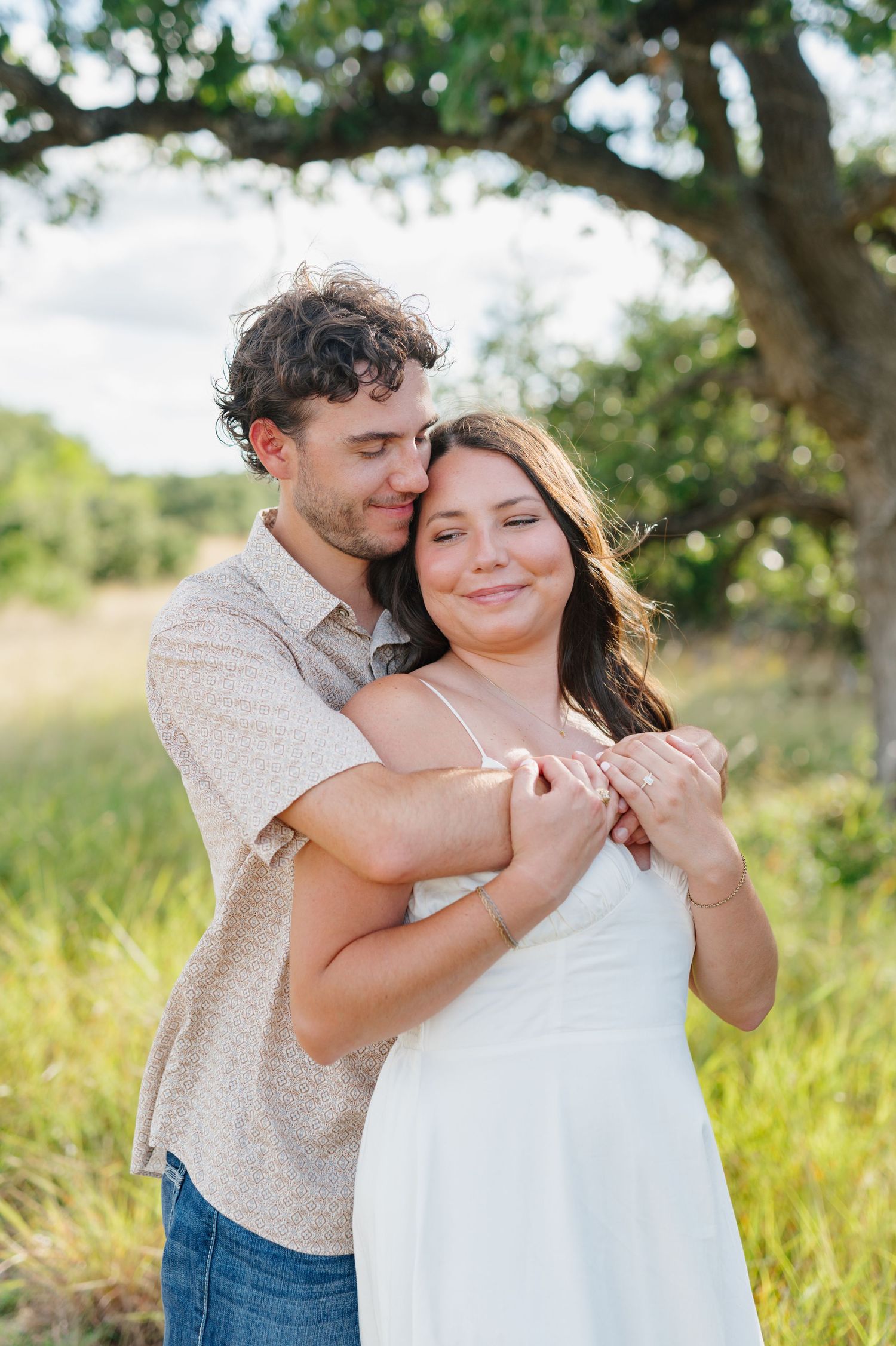 A romantic couple embraces in a sunny meadow with tall grass and trees in the background.
