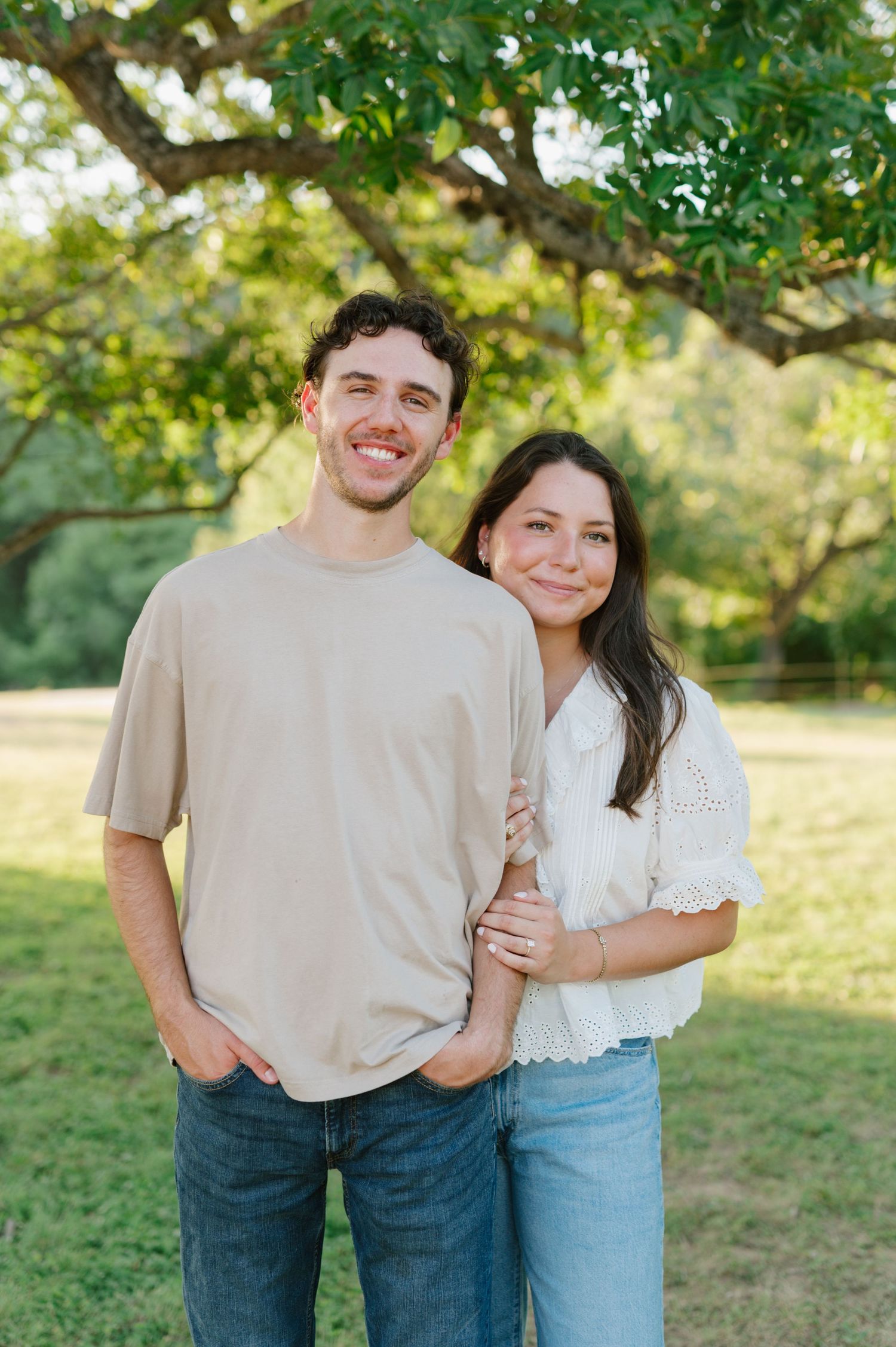 A couple shares sweet candid moments together in a sunny outdoor field with green trees in the background.