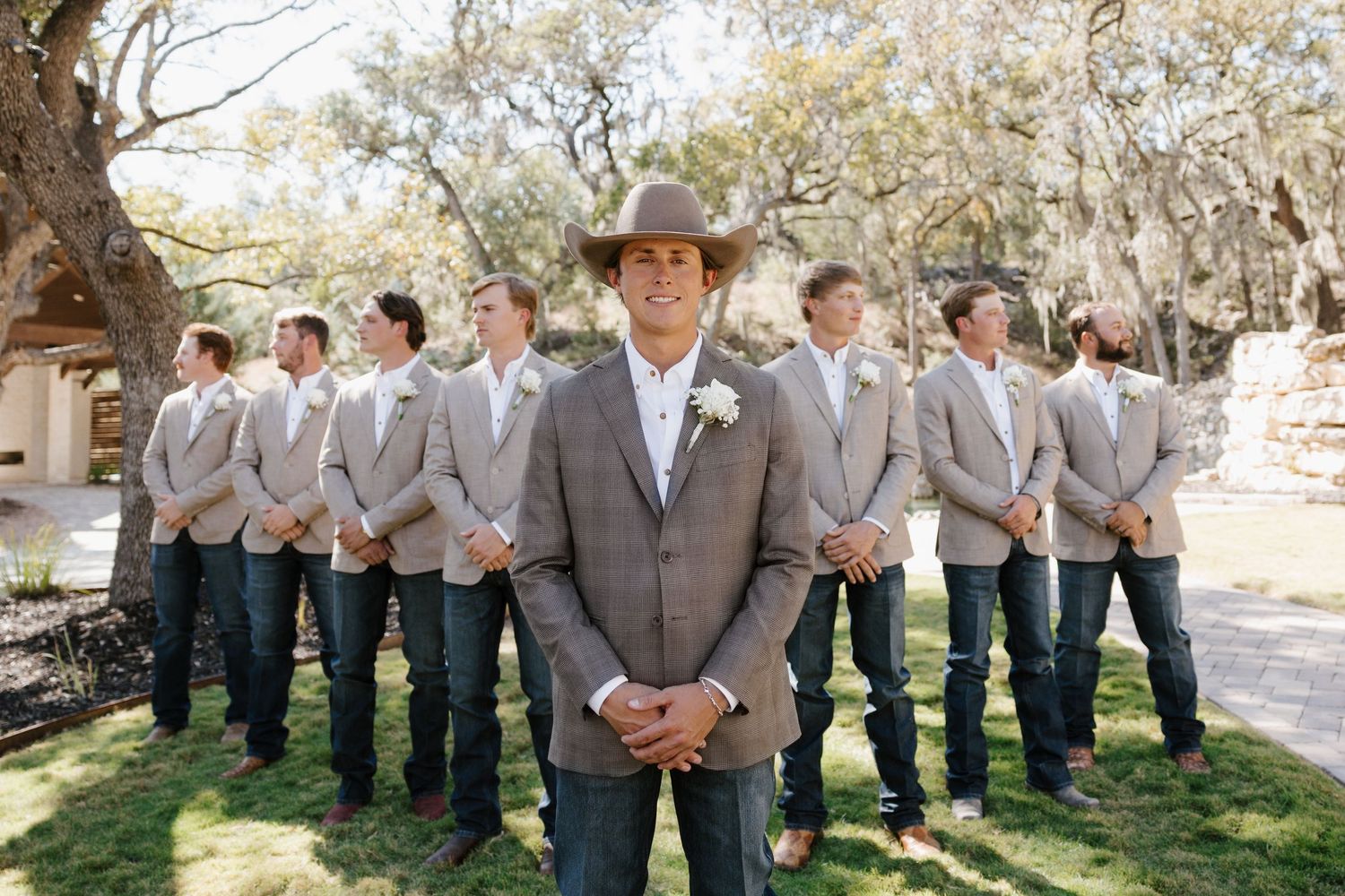 A wedding party in gray suits and brown cowboy boots stands in line outdoors on a sunny day.