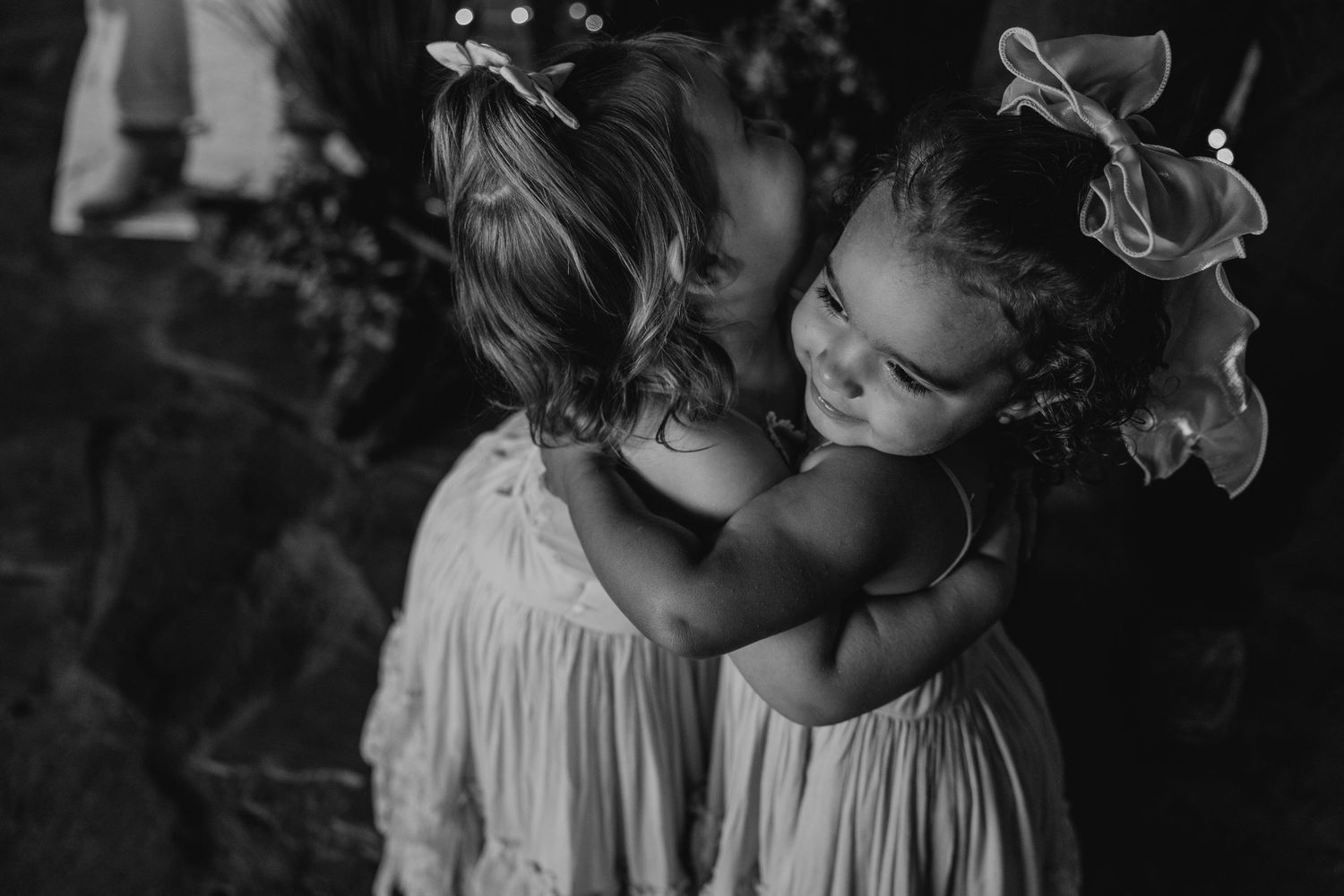 Two young flower girls in white dresses sharing an intimate moment in black and white photography.