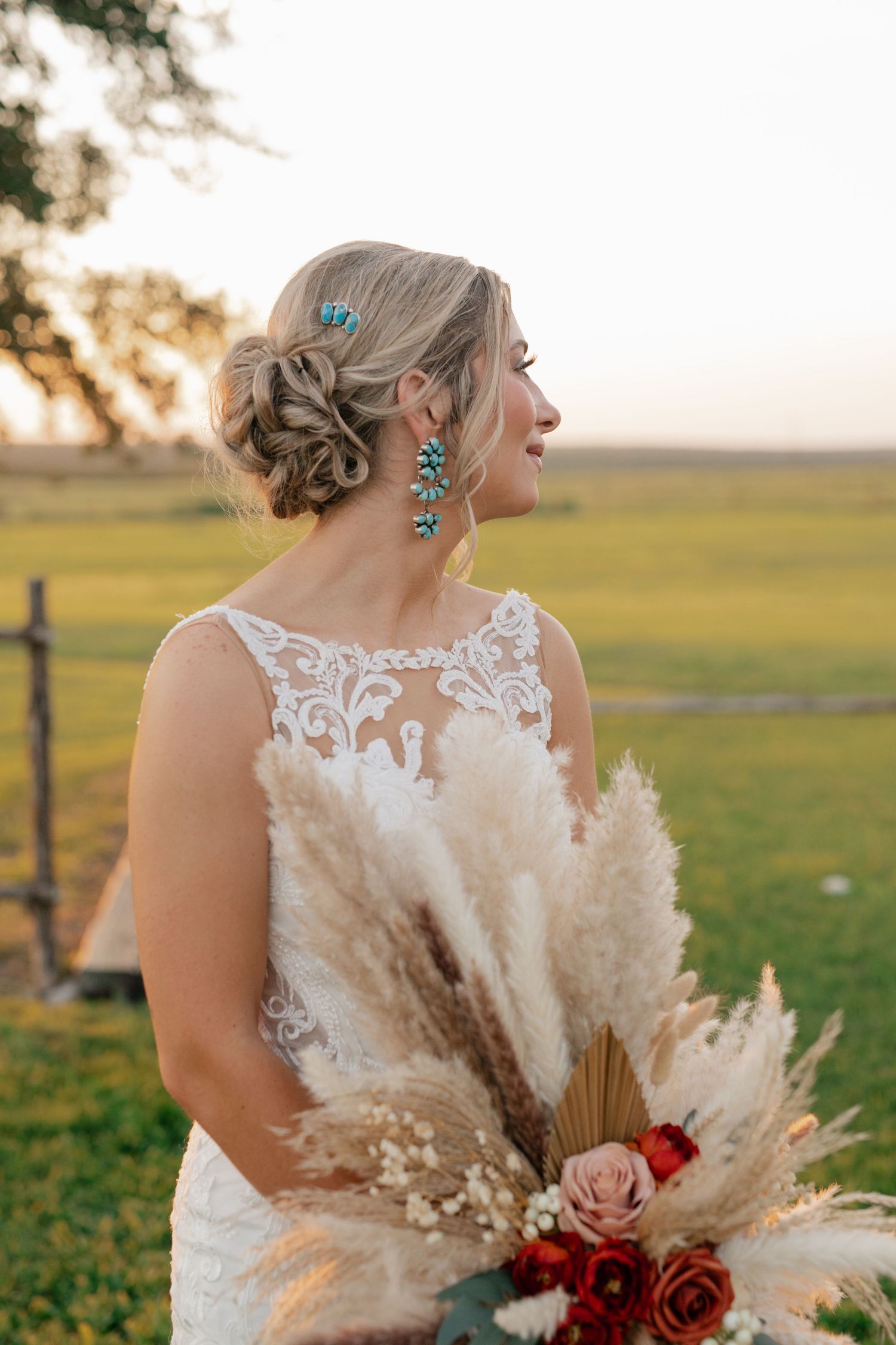 Elegant bride in lace dress holds dried pampas grass and red rose bouquet against sunset field backdrop.