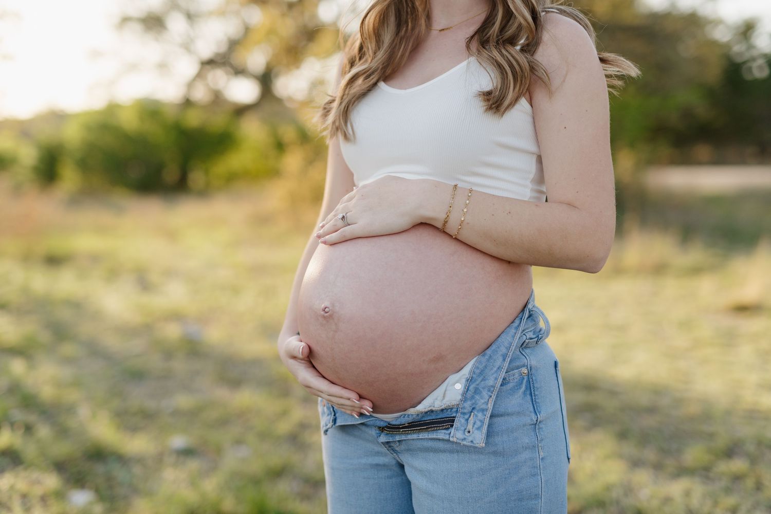 Pregnant woman in white tank top and jeans gently cradling her bare belly in a sunlit outdoor setting.