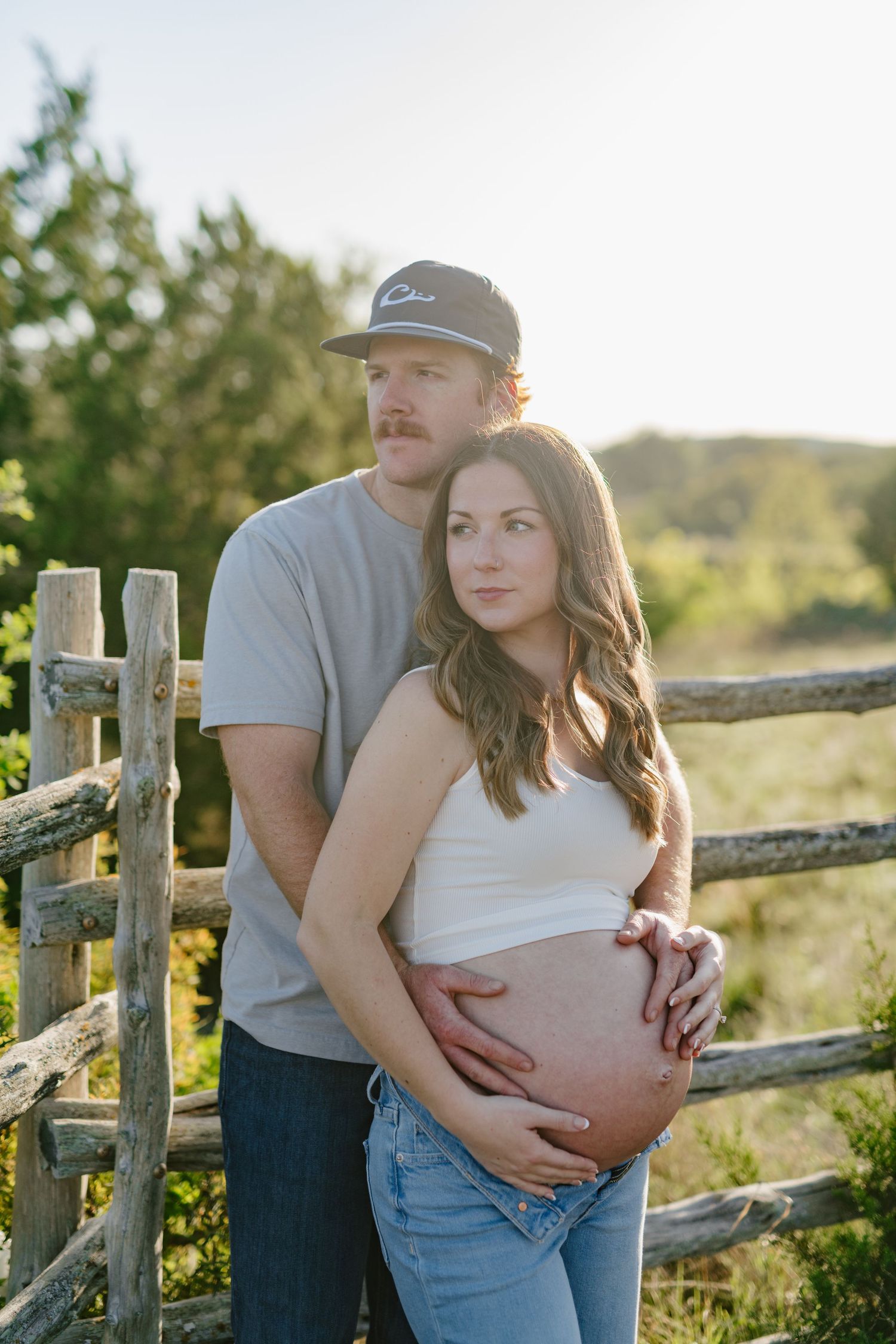 Maternity portrait beside rustic wooden fence during sunset in countryside setting.