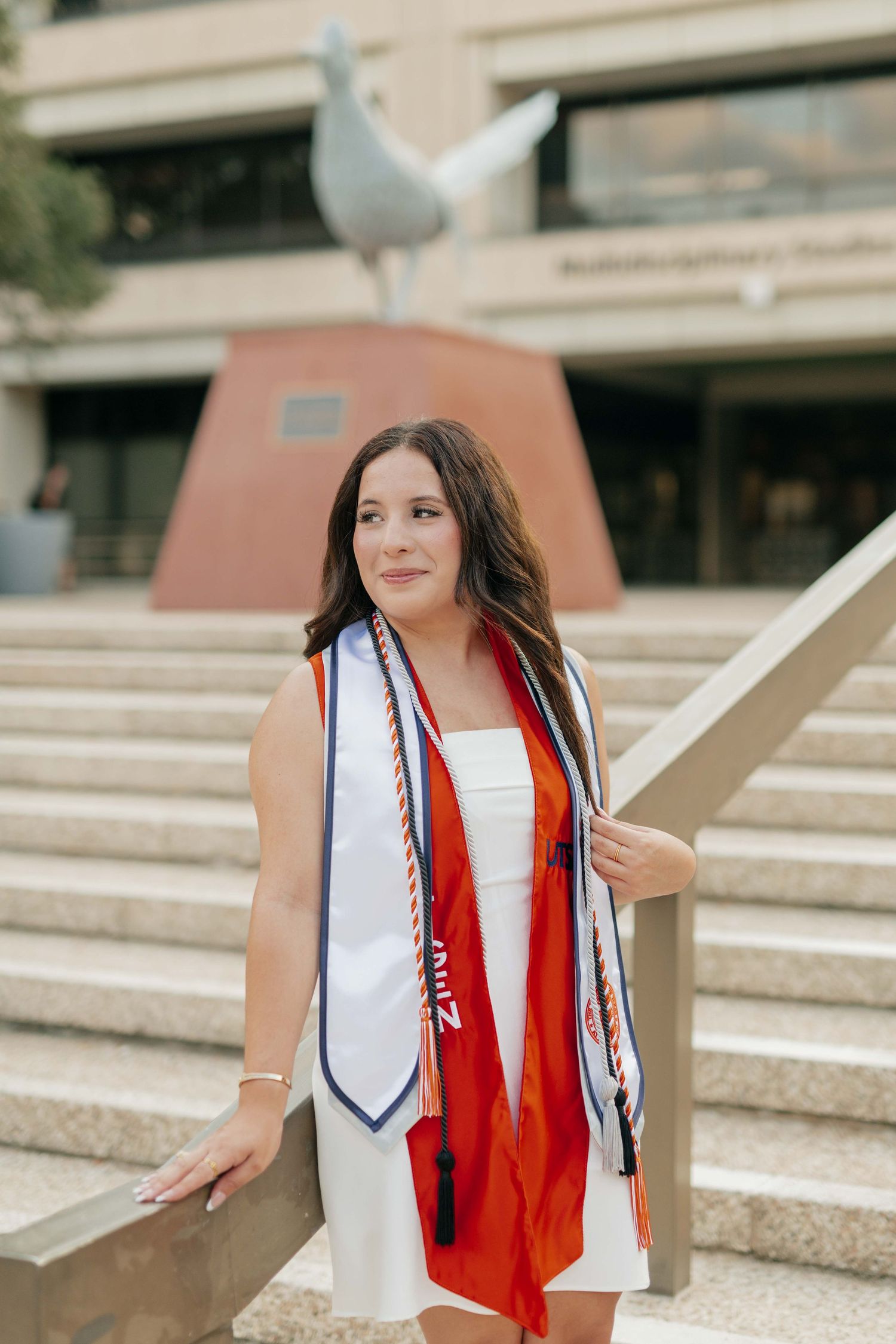 Graduate wearing white dress and graduation stole poses by campus monument.
