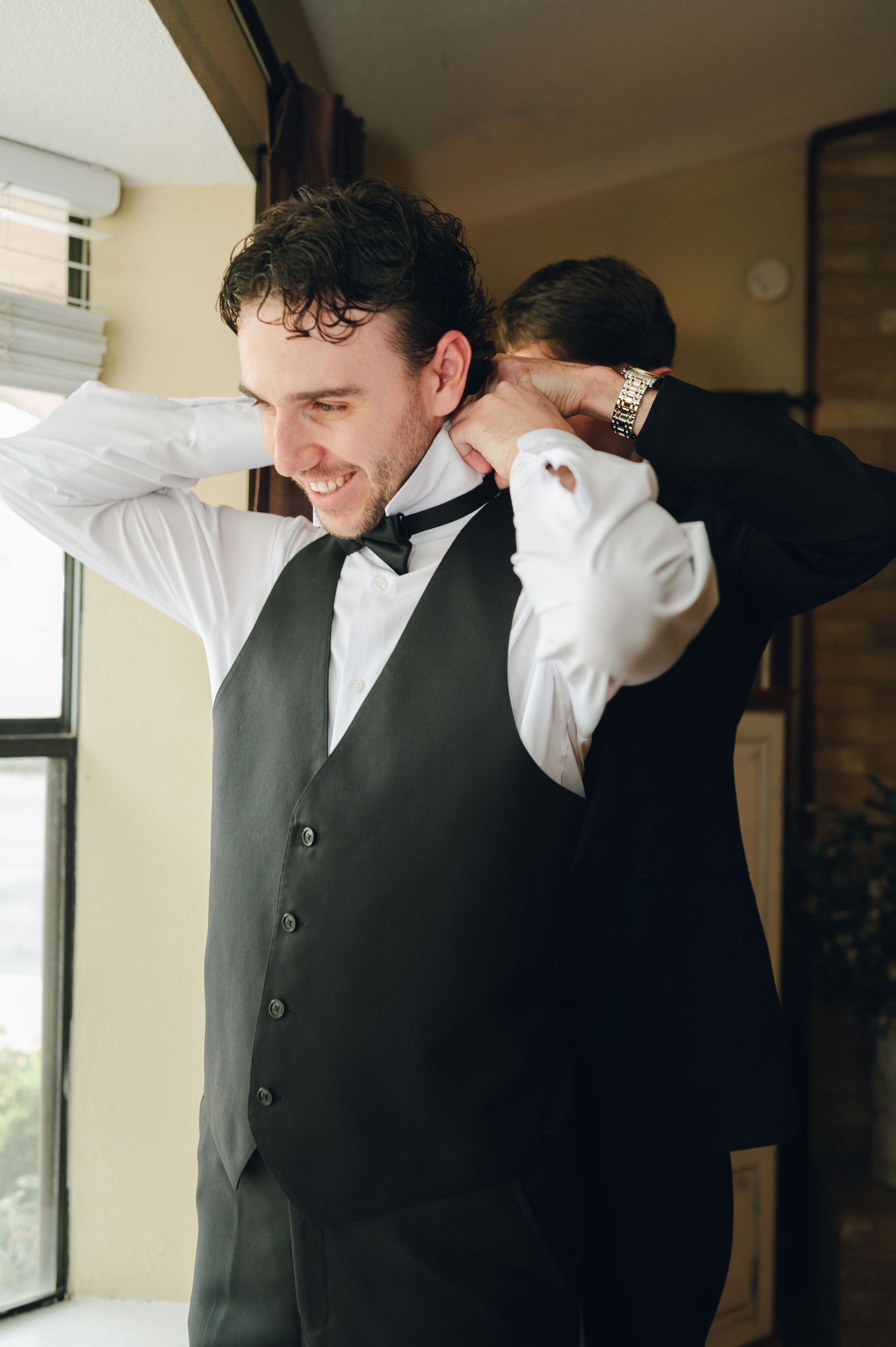 Groom smiling as groomsman helps adjust his black bow tie and tuxedo vest before wedding ceremony.