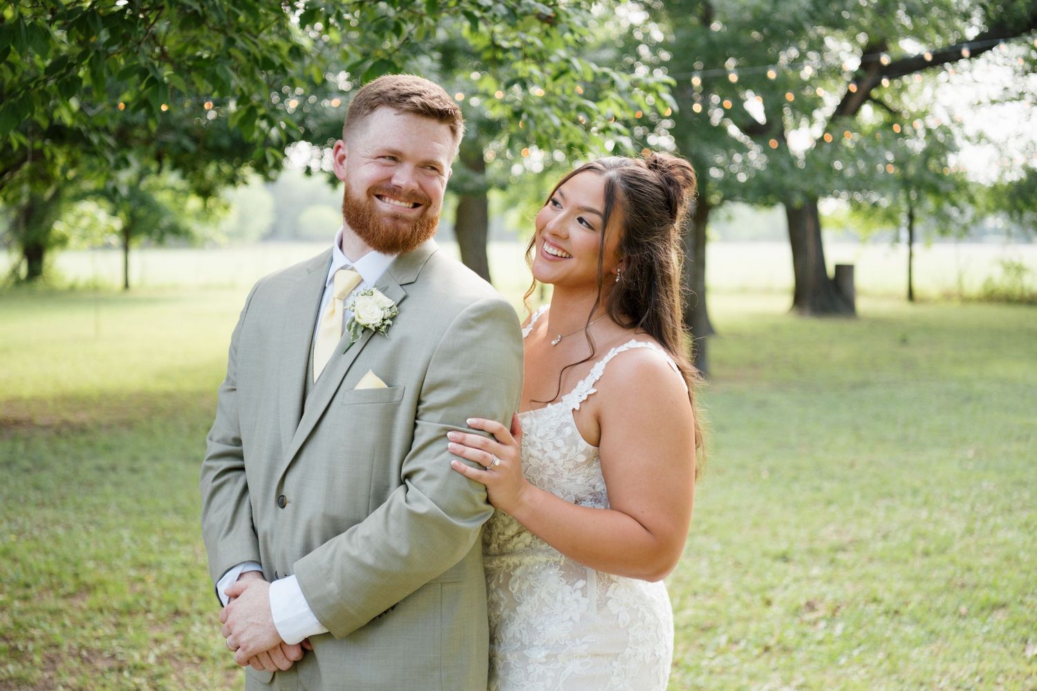 Smiling newlywed couple poses outdoors, groom in sage suit and bride in white lace wedding dress.