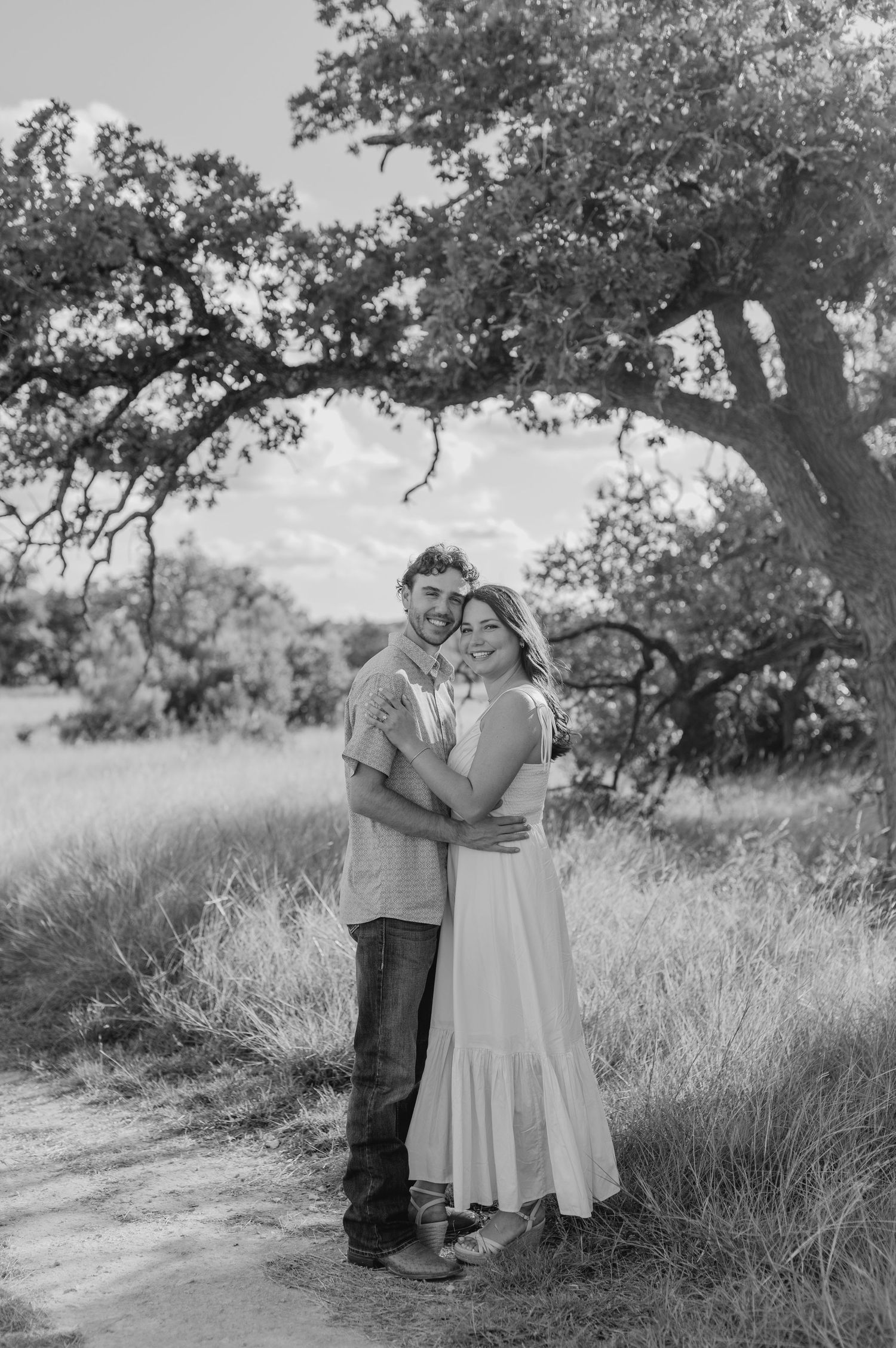 Black and white photo of a couple embracing under a large oak tree in a grassy field.
