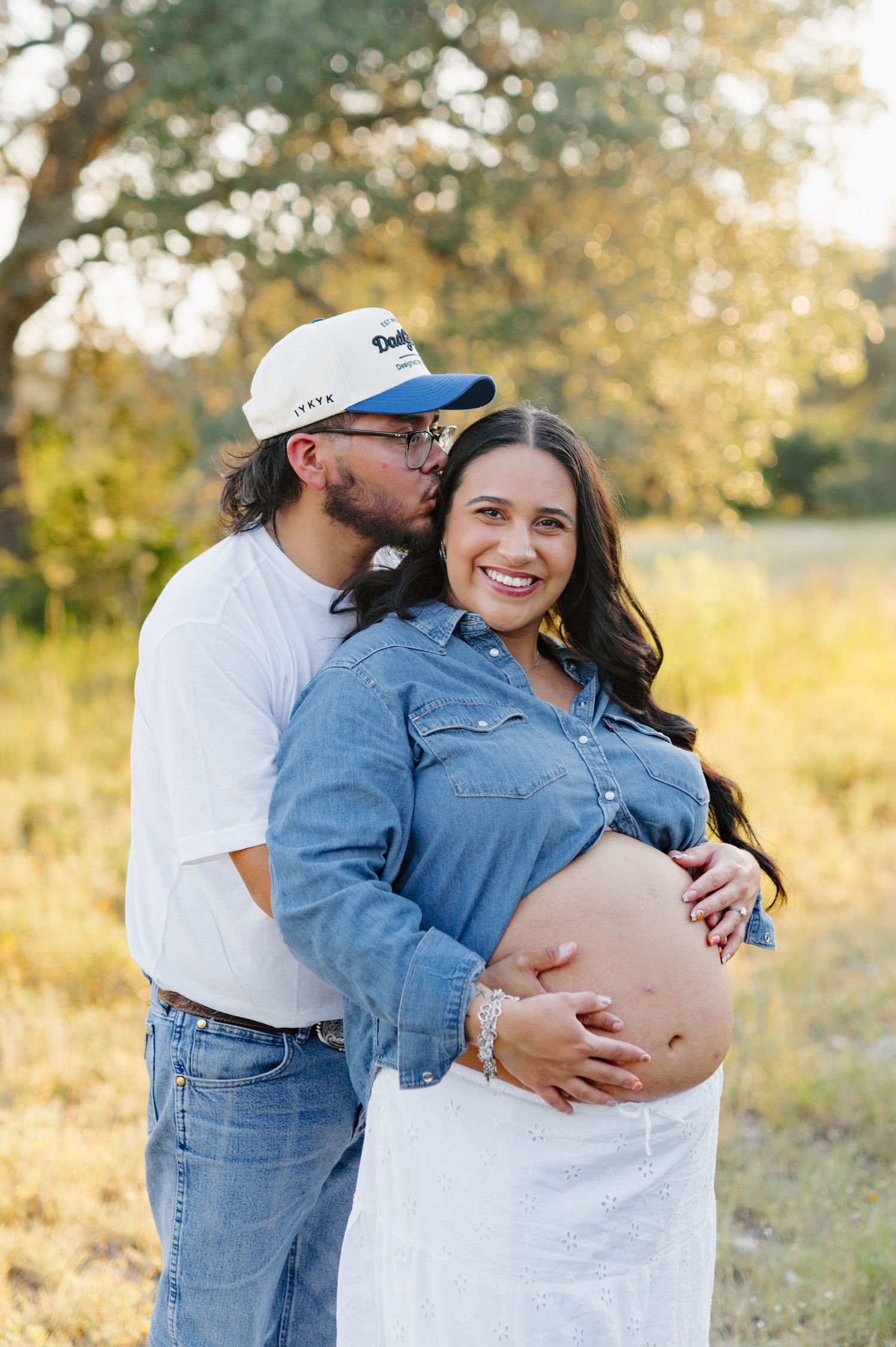 A couple embraces in a sun-drenched field during a maternity photo session wearing denim and white outfits.