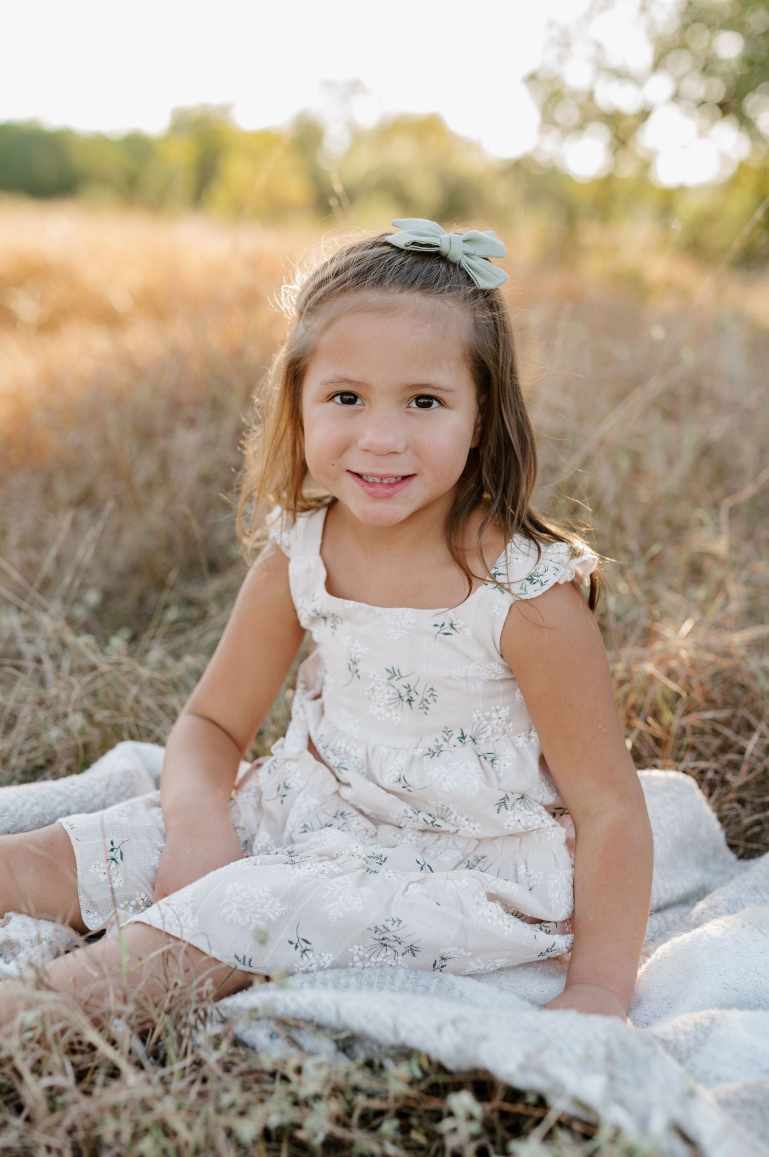 A child in a white floral dress sits on a blanket in a sunlit field during golden hour.