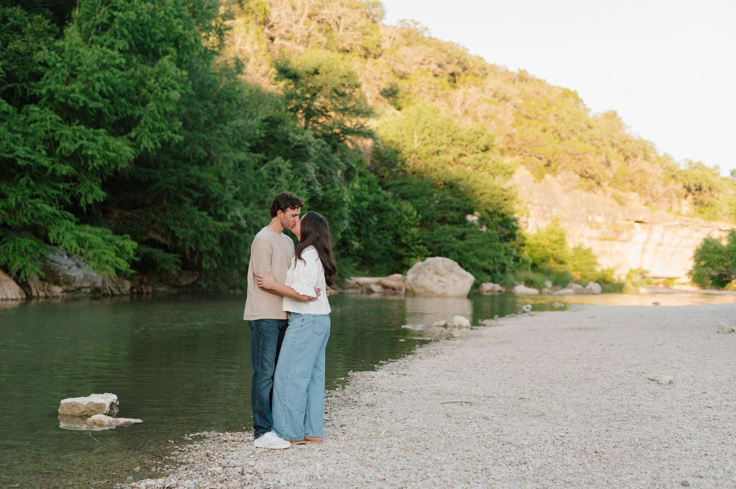 Couple standing by a serene riverside with mountains and greenery in the background.