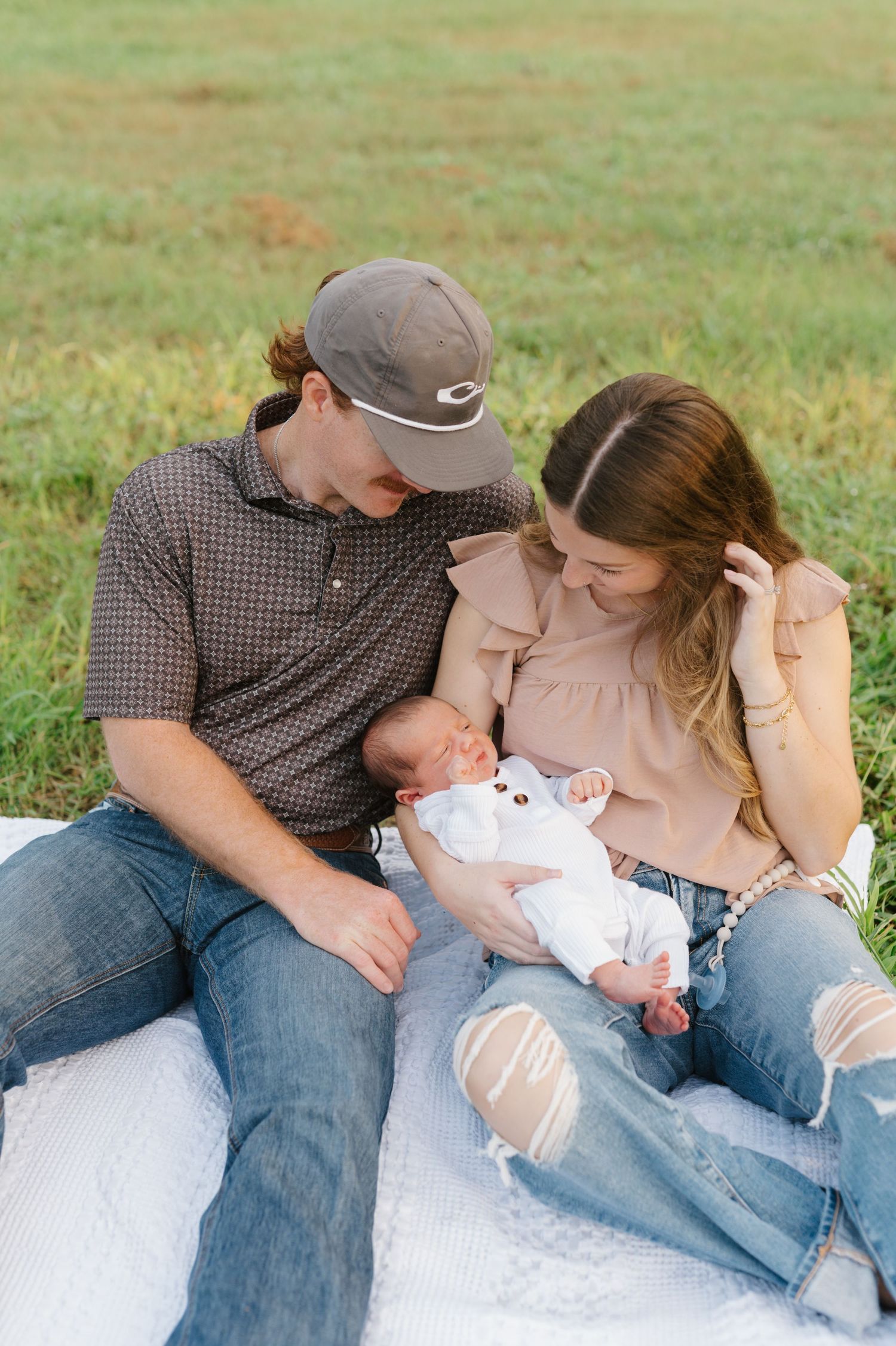 Parents sitting on blanket in grass sharing tender moment with newborn baby wearing white outfit.
