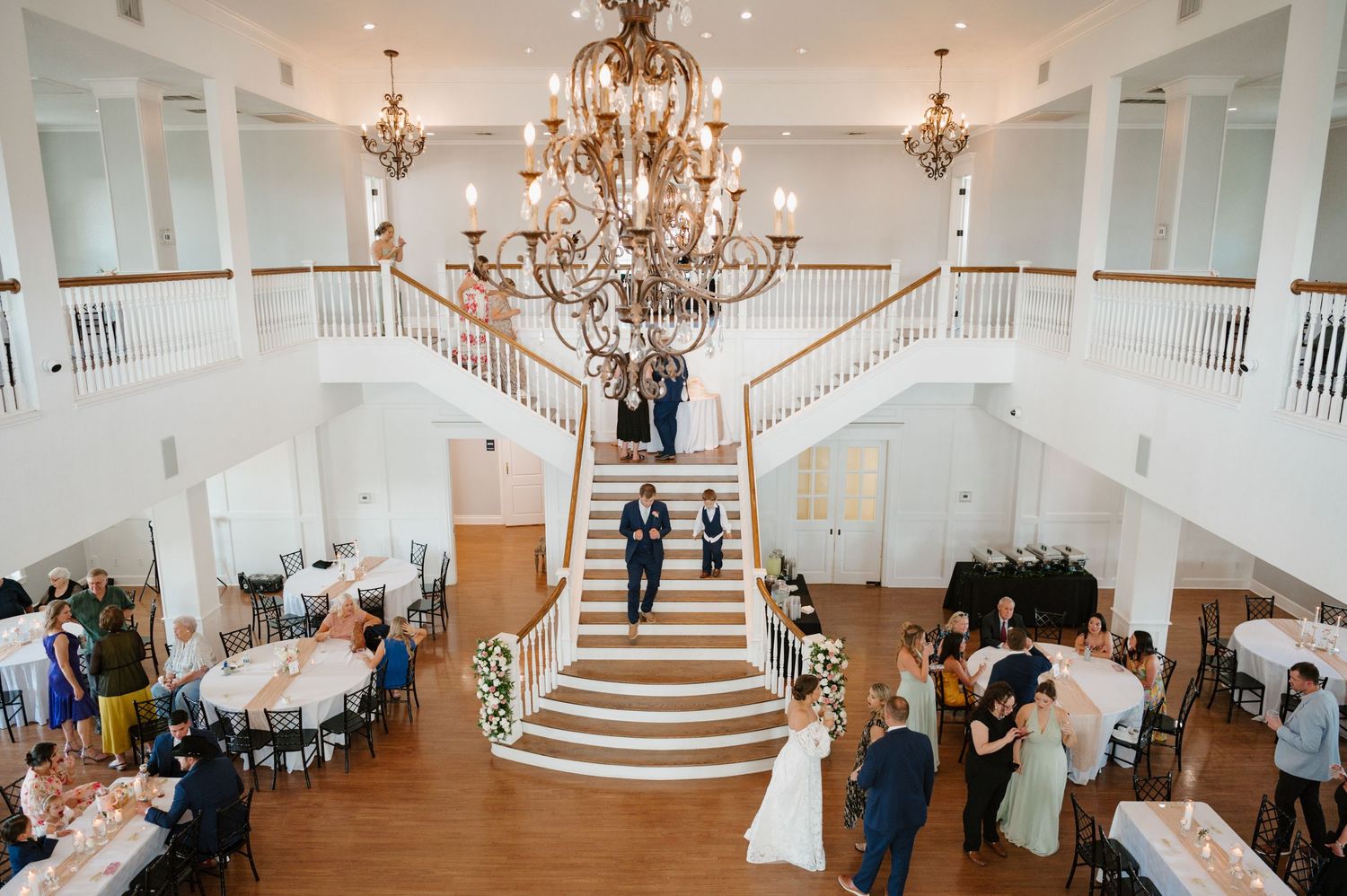 Grand white ballroom with elegant chandelier and sweeping staircase during wedding reception.