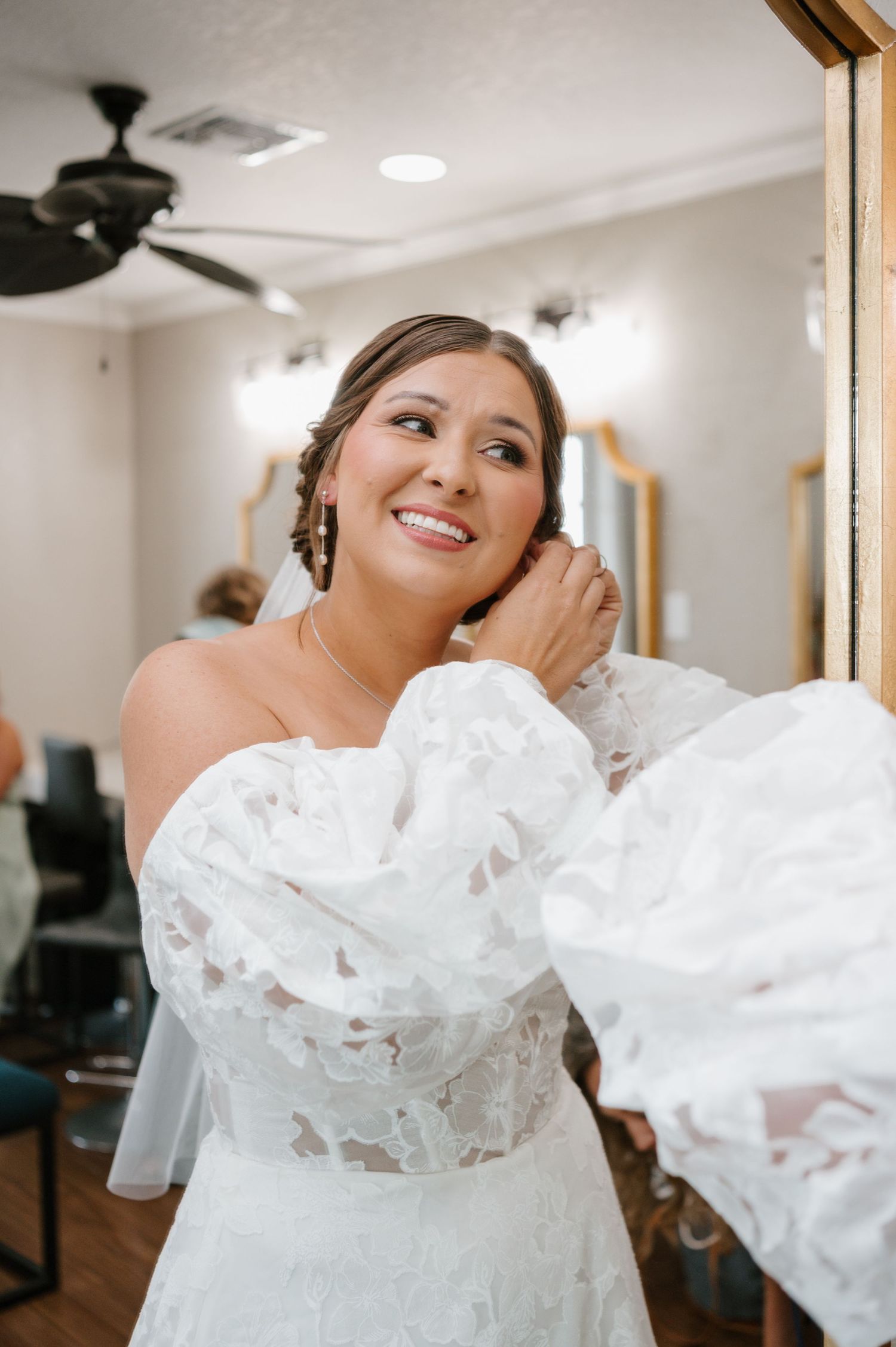 A bride in a flowing white wedding dress looks cheerful while getting ready in a room.
