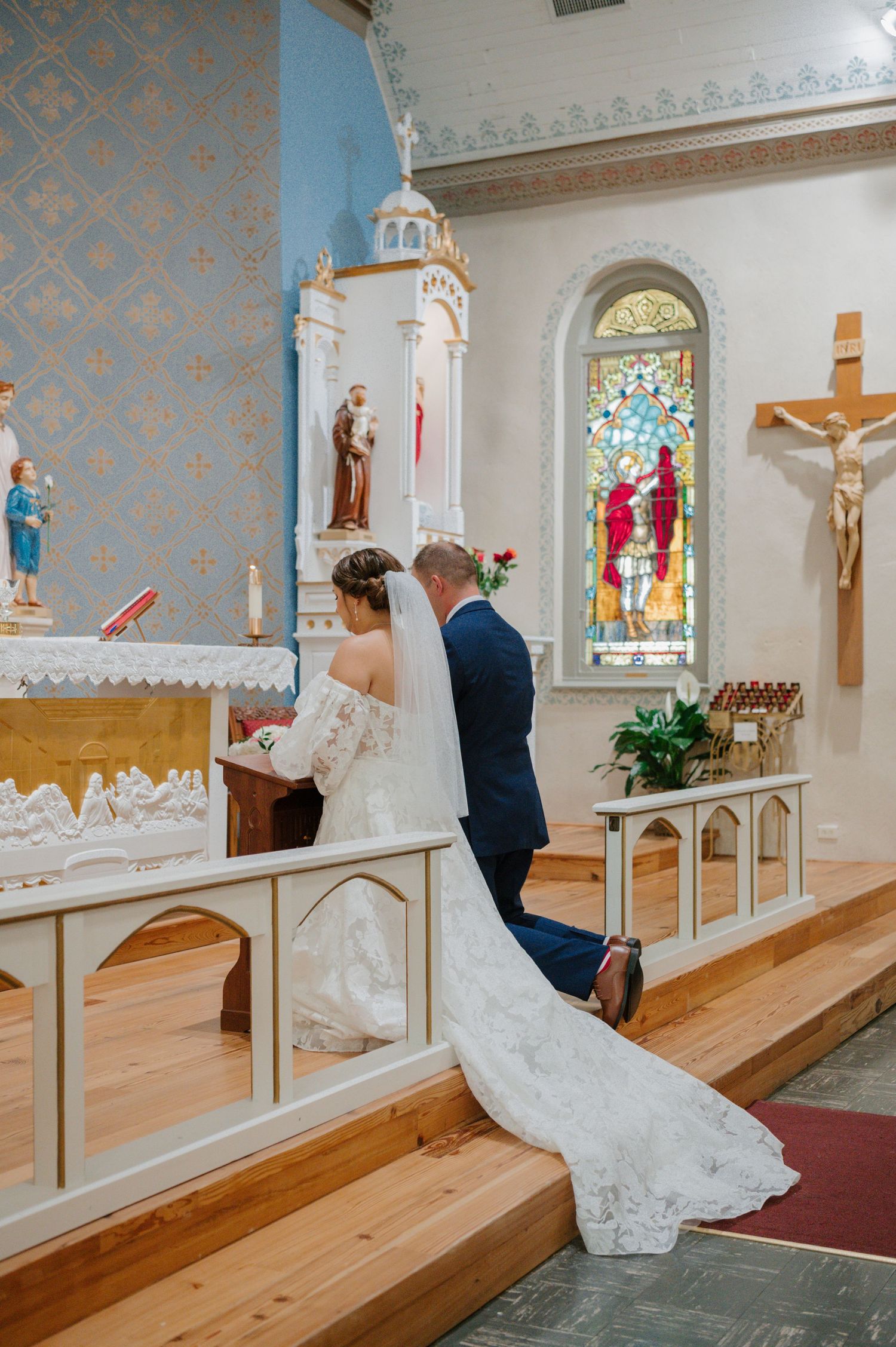 Bride in lace gown and groom pray together inside ornate Catholic church with stained glass windows.