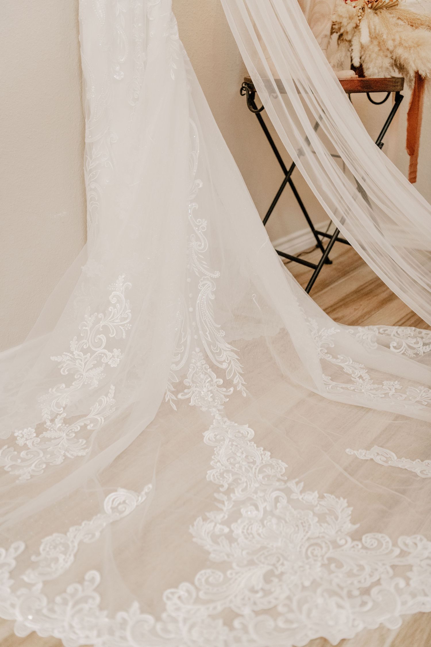 Close-up detail of delicate white lace and embroidery on a flowing wedding dress train against a light background.