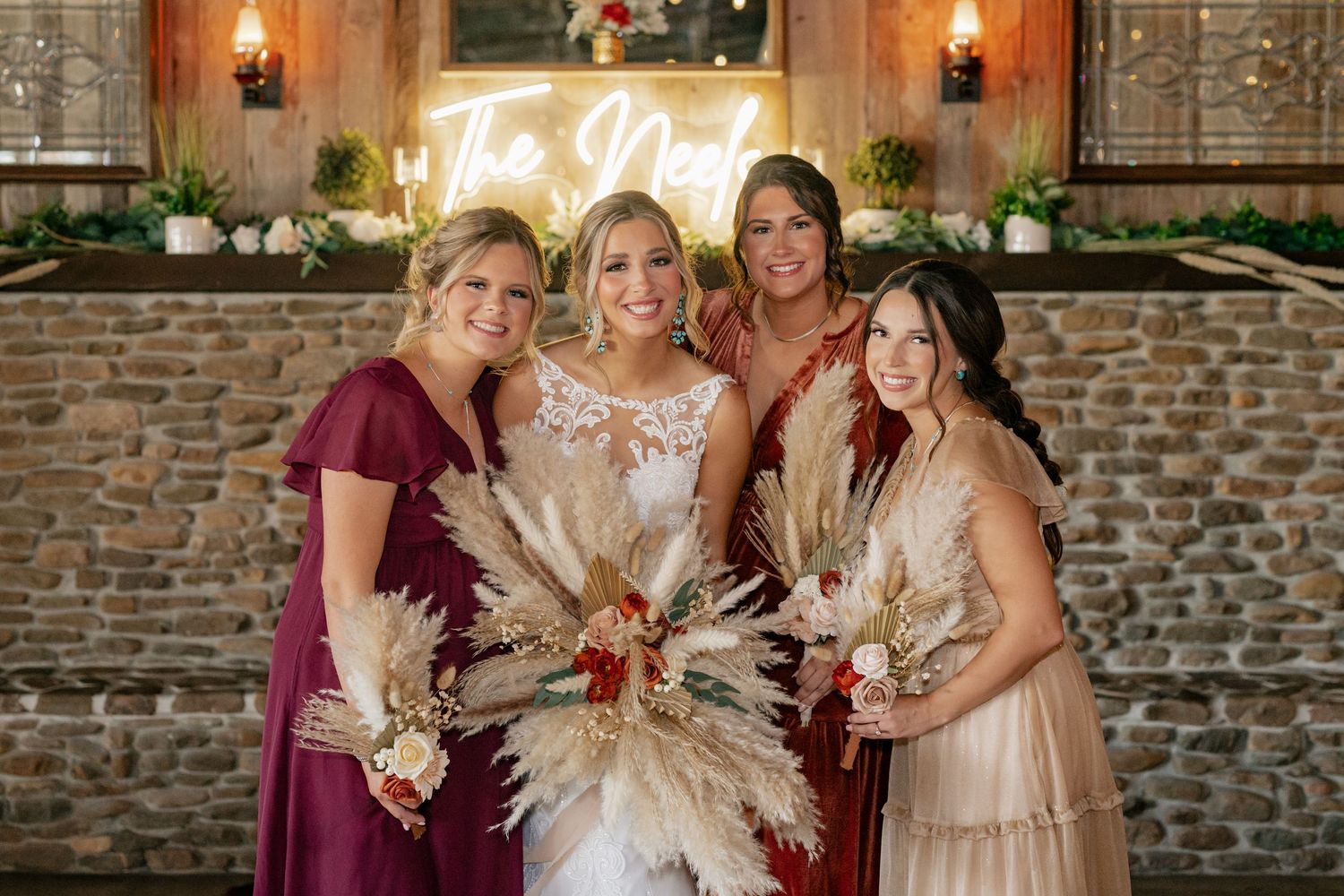 A wedding party posing together with pampas grass bouquets against a stone wall backdrop.
