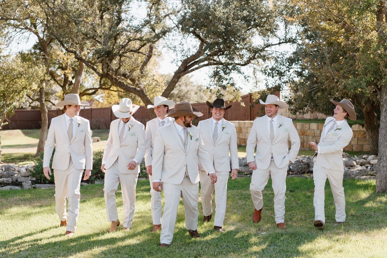 Wedding party wearing crisp white suits walking across a sunlit garden lawn beneath large trees.