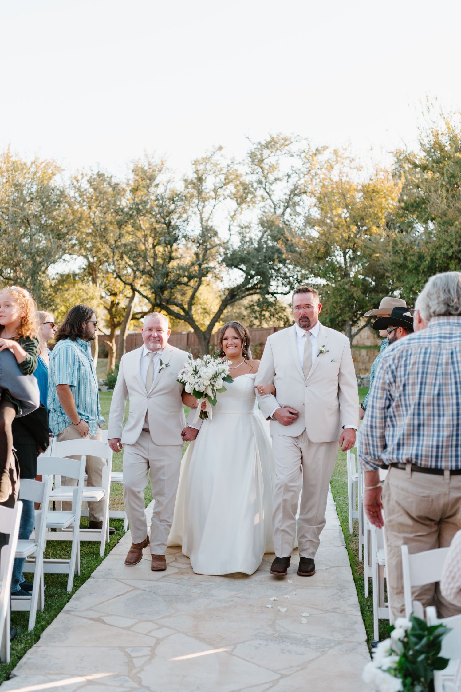 A bride and groom walk down an outdoor stone pathway lined with white chairs and trees during a garden ceremony.