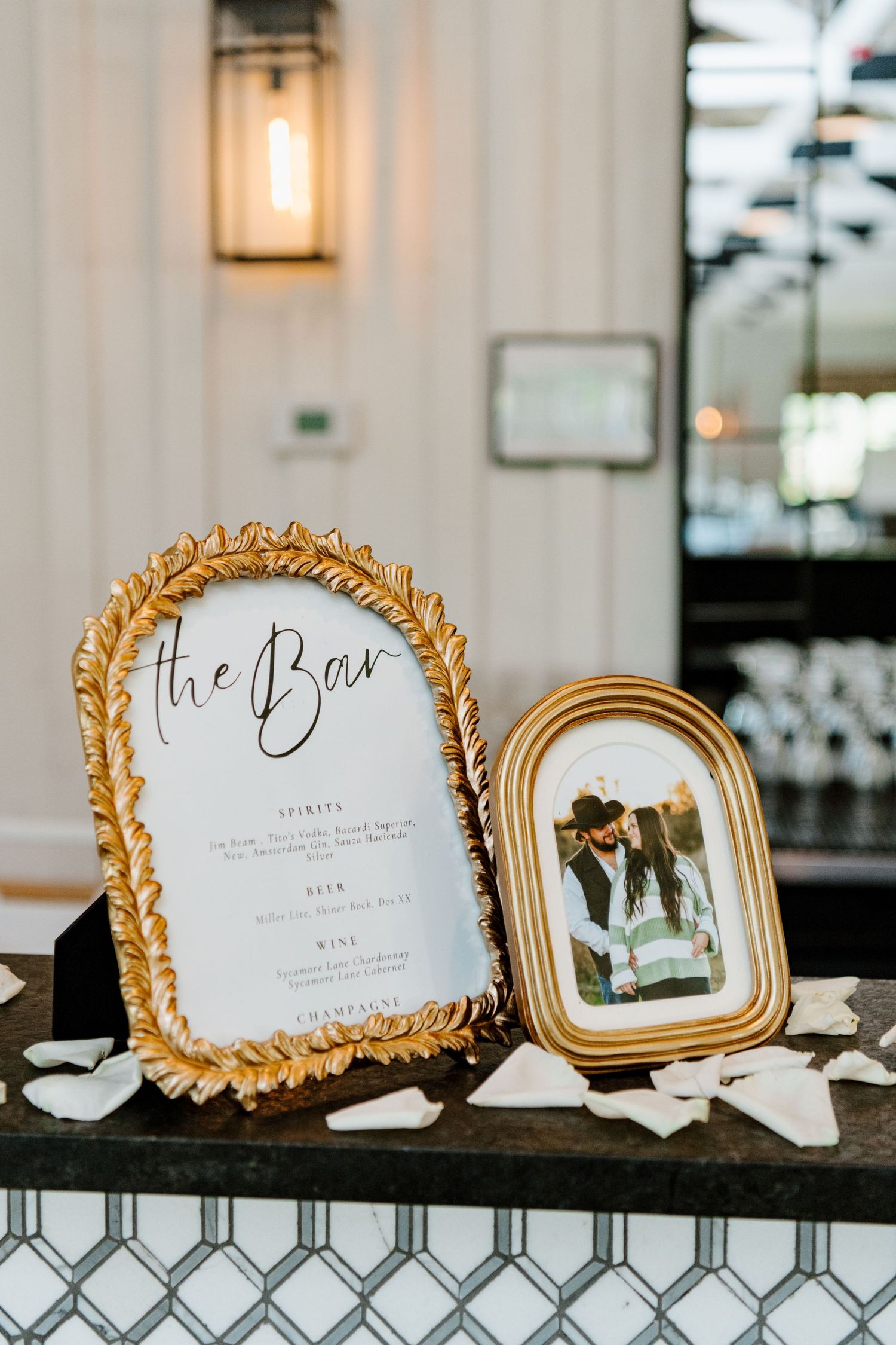 Elegant gold-framed bar menu and decorative photo frame displayed on dark surface with scattered white petals.