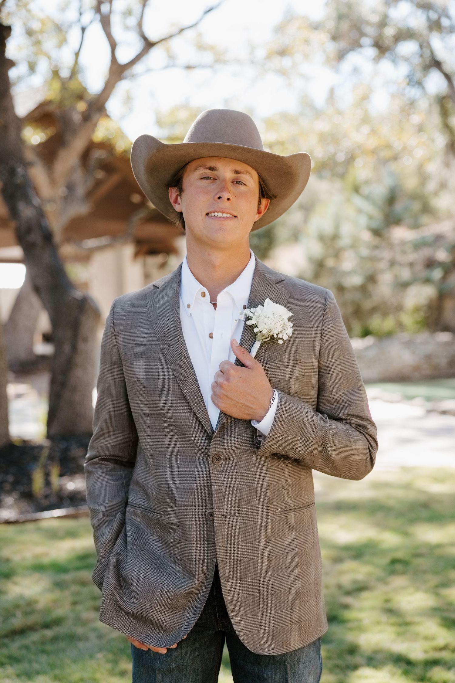 Groom in a gray suit and cowboy hat poses outdoors in a rustic setting.