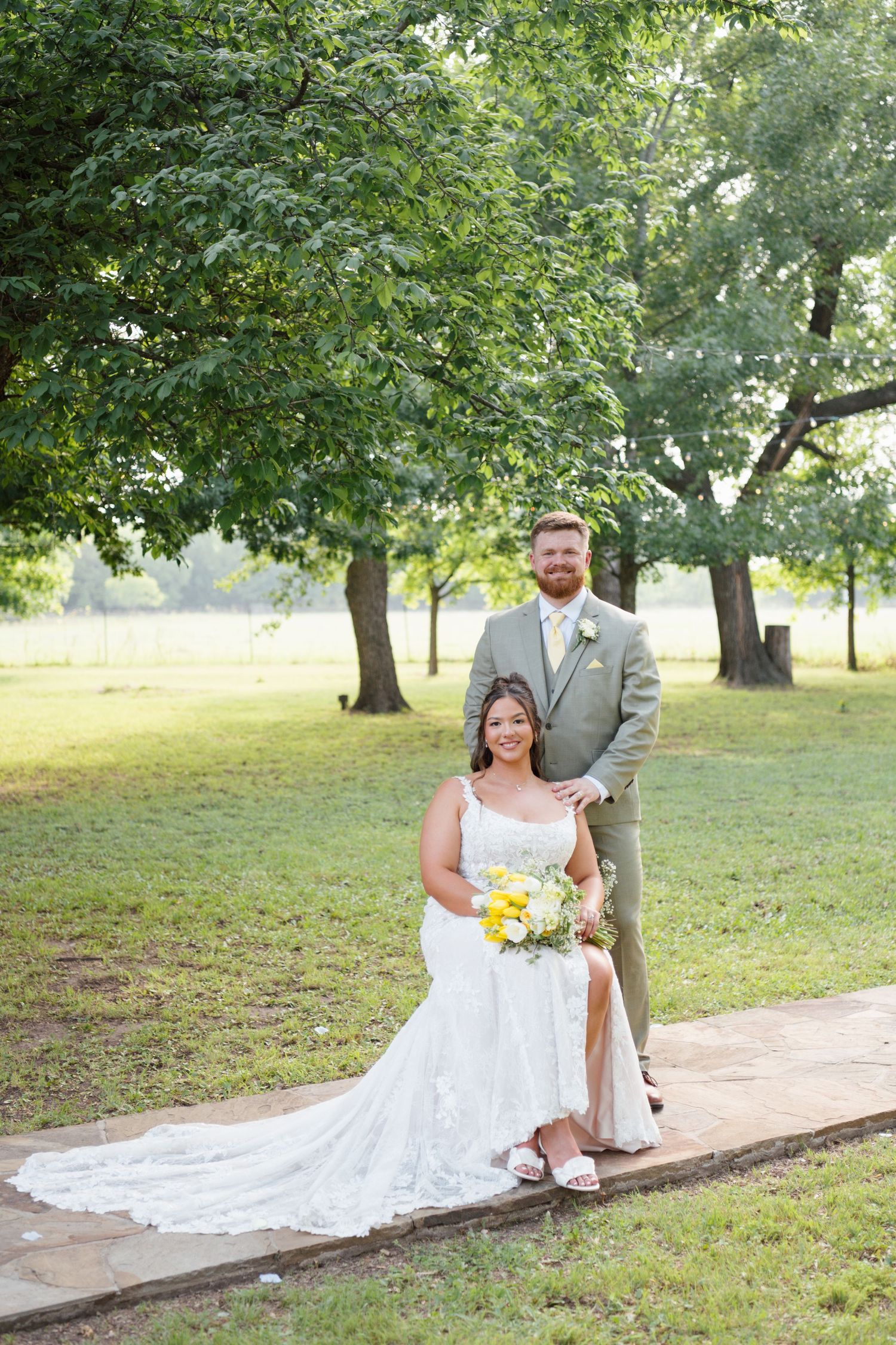 Bride seated in white lace gown holding yellow bouquet with groom in tan suit standing behind her outdoors.